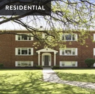 A three-story brick residential building with white window frames and a central entrance, surrounded by a grassy lawn and trees.
