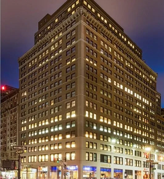 Night view of a tall, multi-story office building with illuminated windows, located at a city intersection with street signs and traffic lights.
