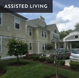 Assisted living apartment complex with parked cars and landscaped front yard, under a partly cloudy sky.