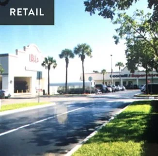 Street view of a retail shopping area with stores, palm trees, and parking lot.