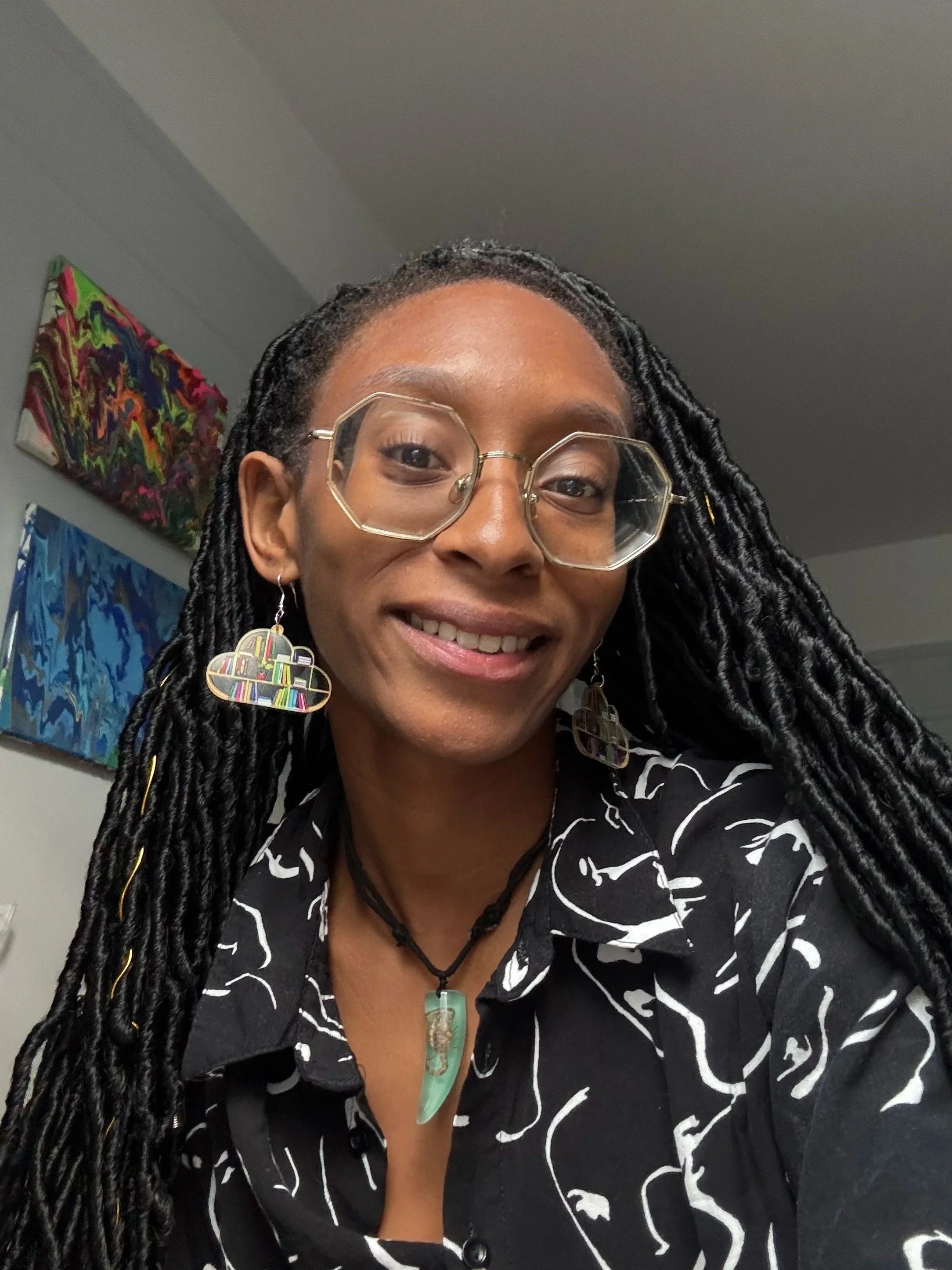 A woman with long dreadlocks wearing large geometric glasses, earrings, a necklace, and a black shirt with white lining, smiling at the camera. Background includes colorful abstract paintings on a wall.