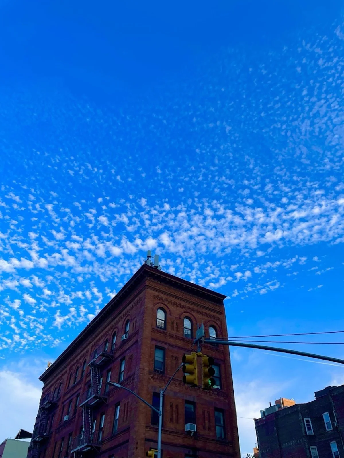 A brick building with fire escapes and windows, under a bright blue sky with scattered white clouds, and traffic signals in the foreground.