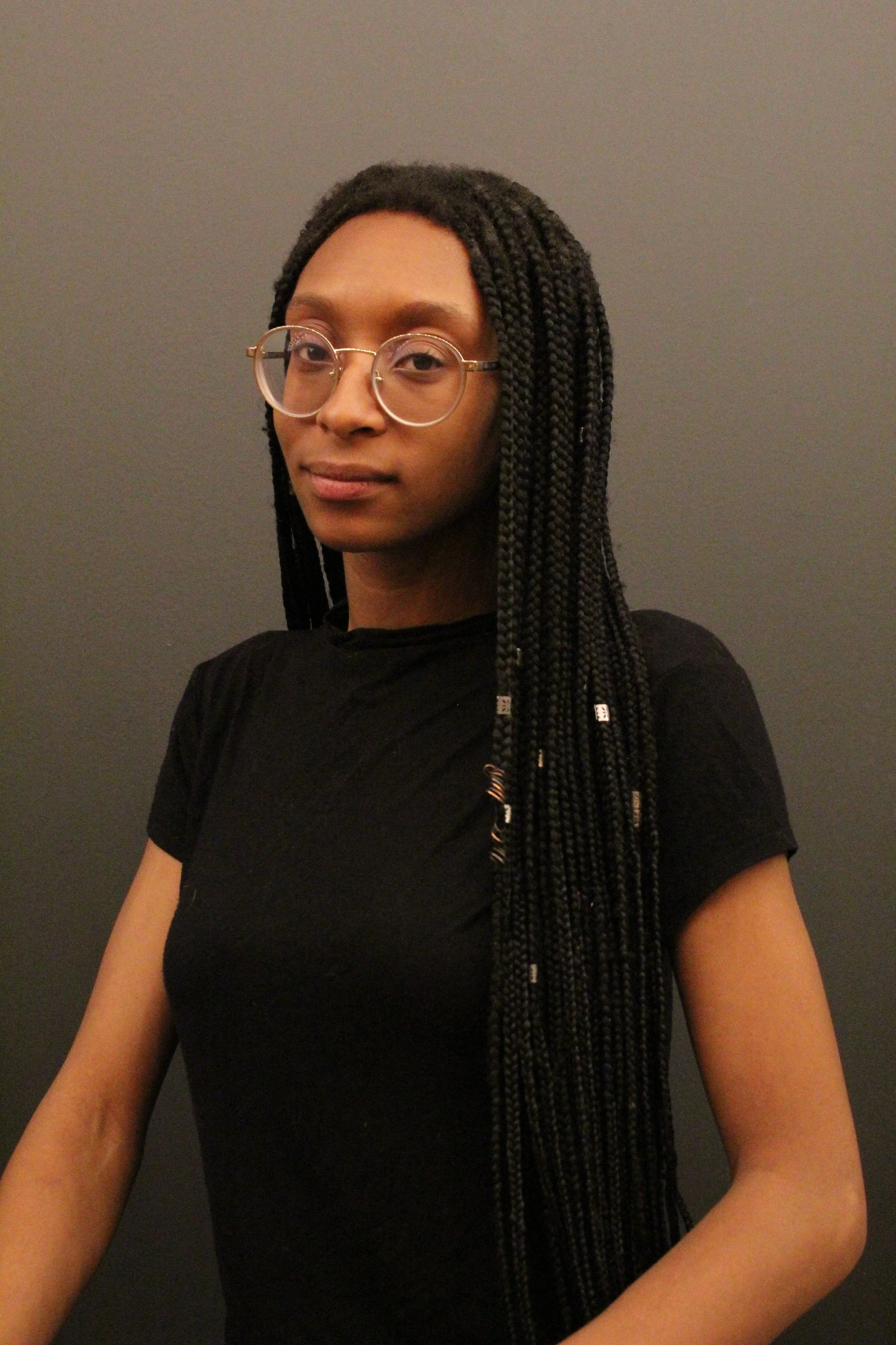 Portrait of Lailani Gibson wearing glasses and long braids against a gray background.