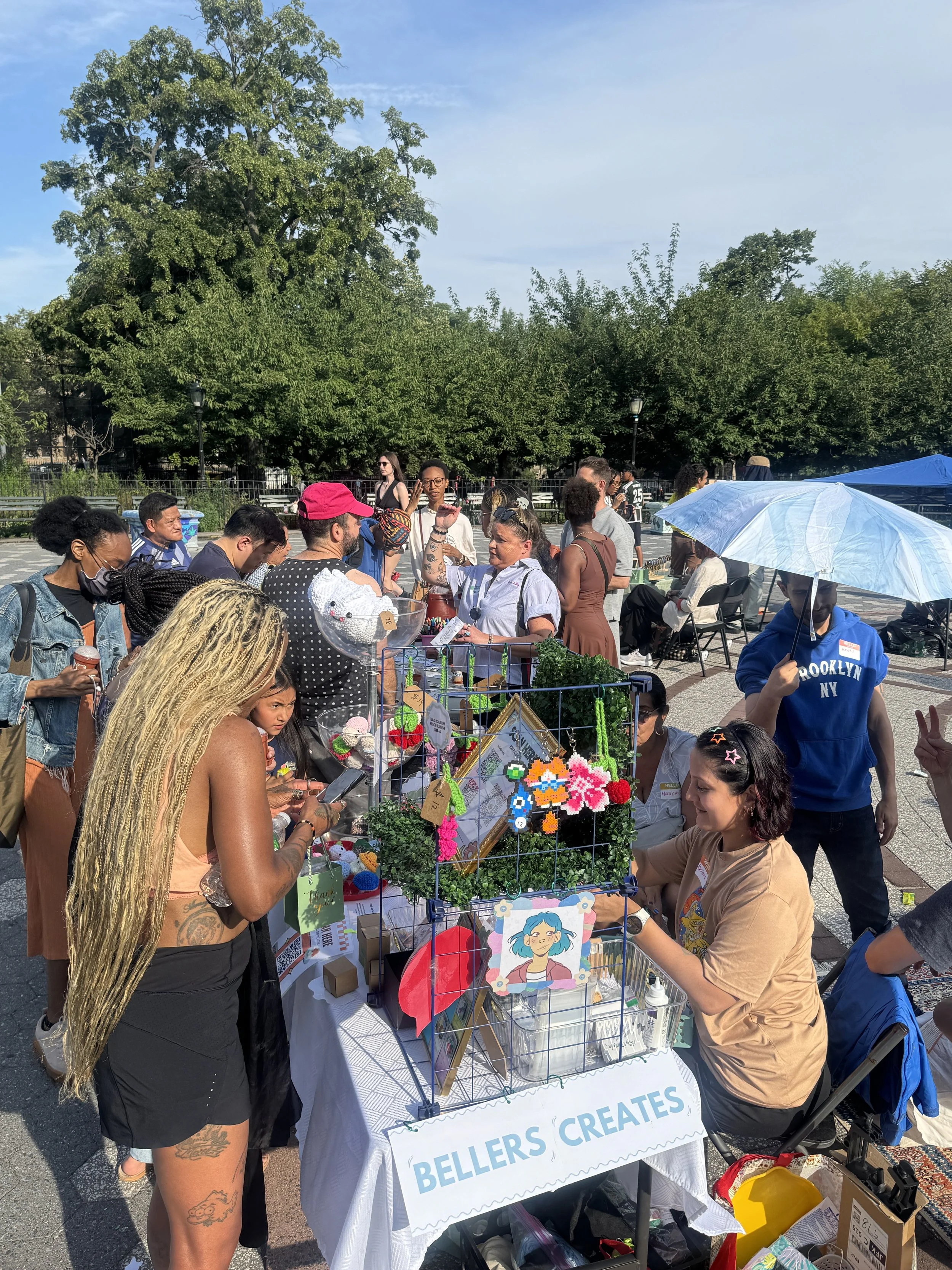 People browsing and shopping at an outdoor street market, with a table displaying DIY crafts, decorations, and artwork, under sunny weather with green trees in the background.