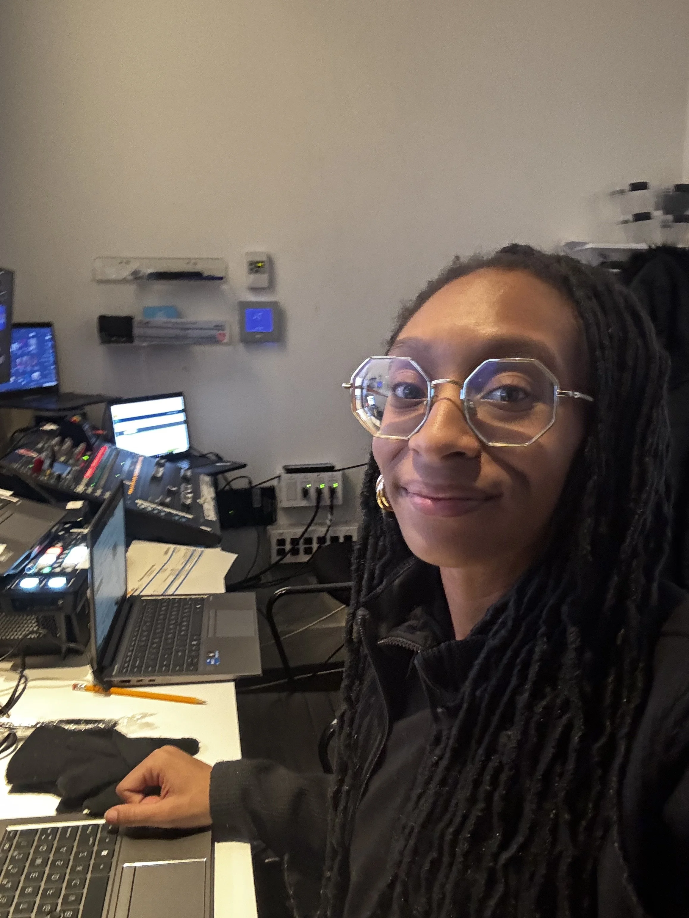A woman with glasses and dreadlocks sitting at a desk with multiple laptops, microphones, and electronic equipment in a workspace or studio.