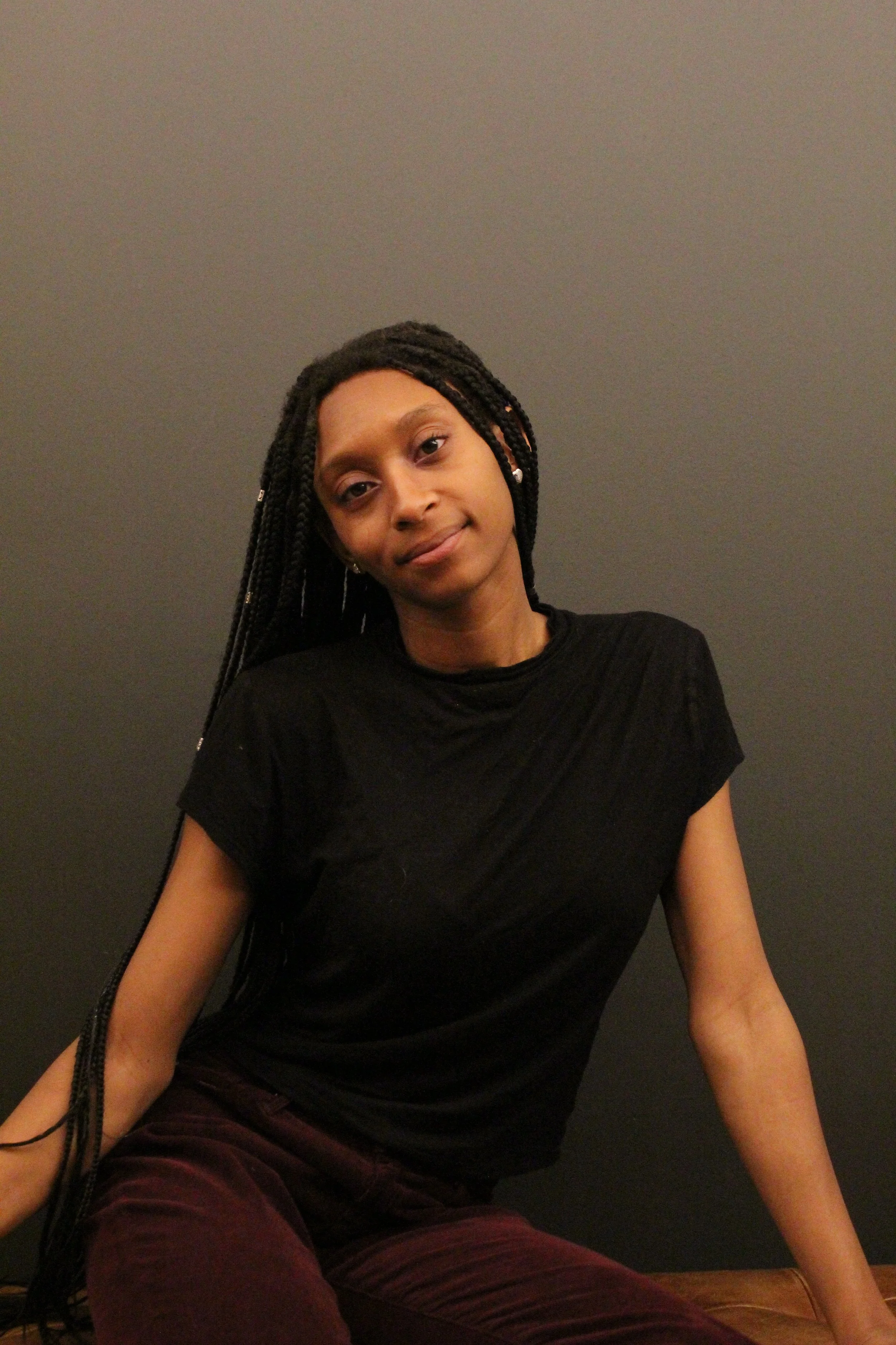 A young woman with braided hair wearing a black shirt and maroon pants, sitting against a plain dark wall.