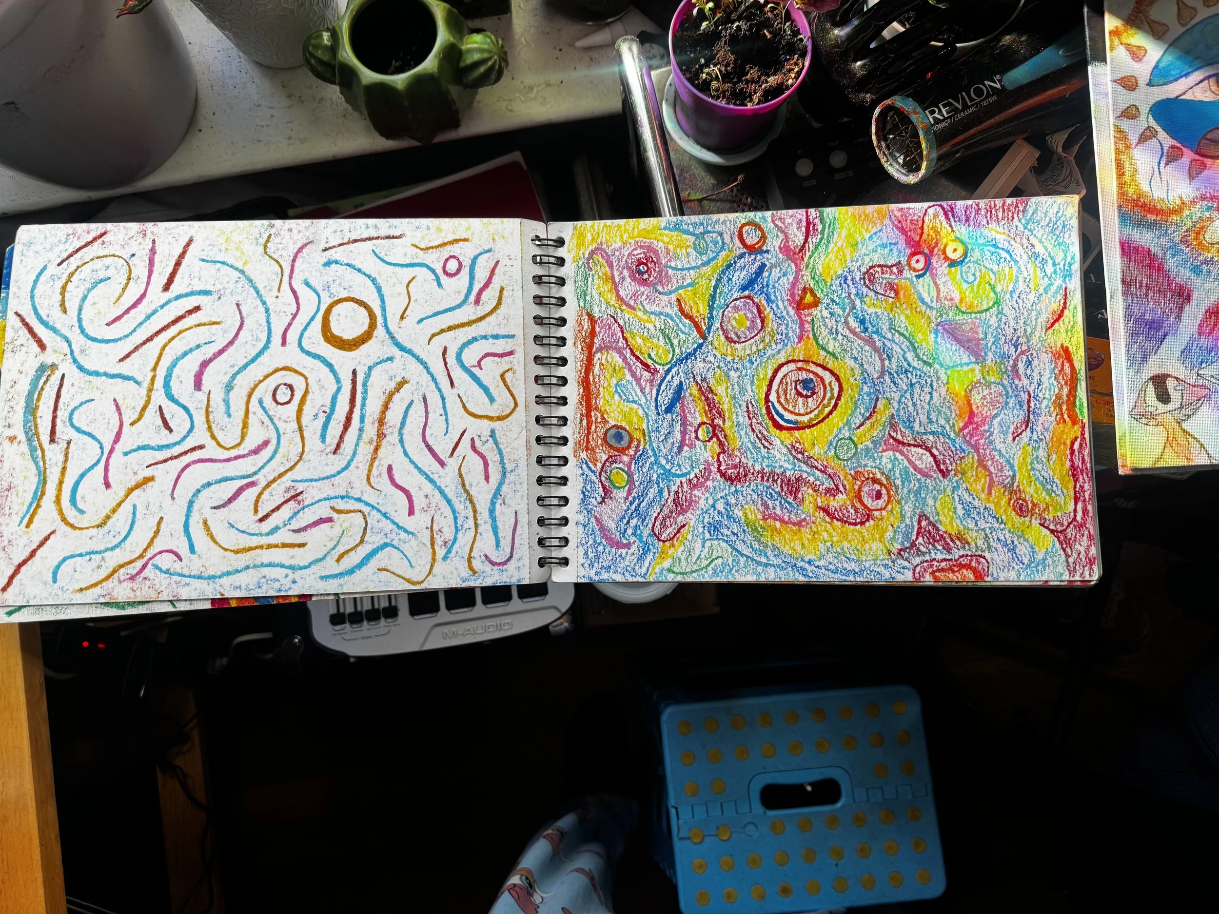 Open sketchbook with colorful, abstract crayon drawings on two pages, situated on a cluttered desk with various art supplies, cups, and a colorful canvas in the background.