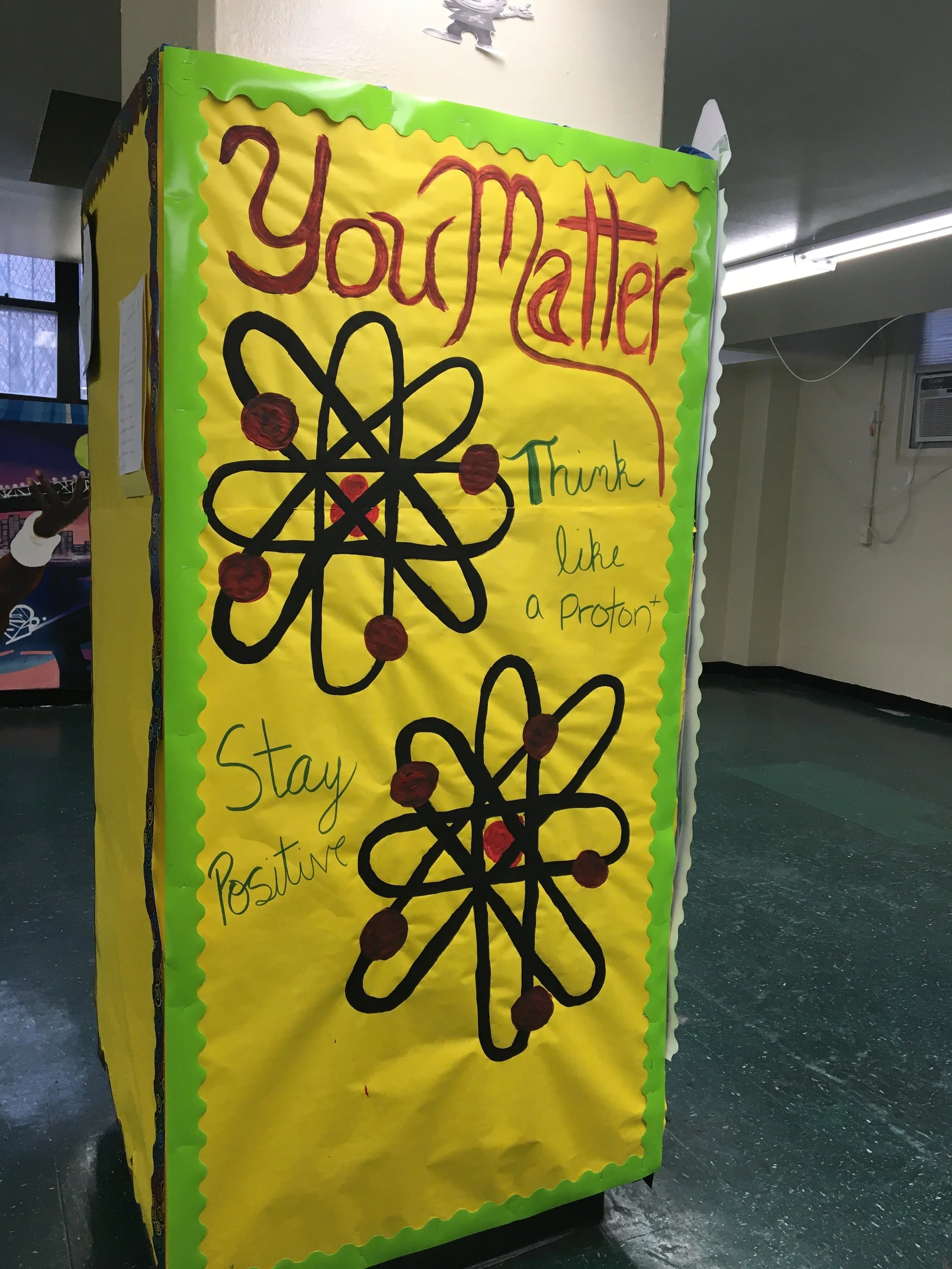 A large yellow poster with a green border that says 'You Matter' in red writing, and 'Think like a Proton' in green, with a black atom illustration and red dots surrounding it.