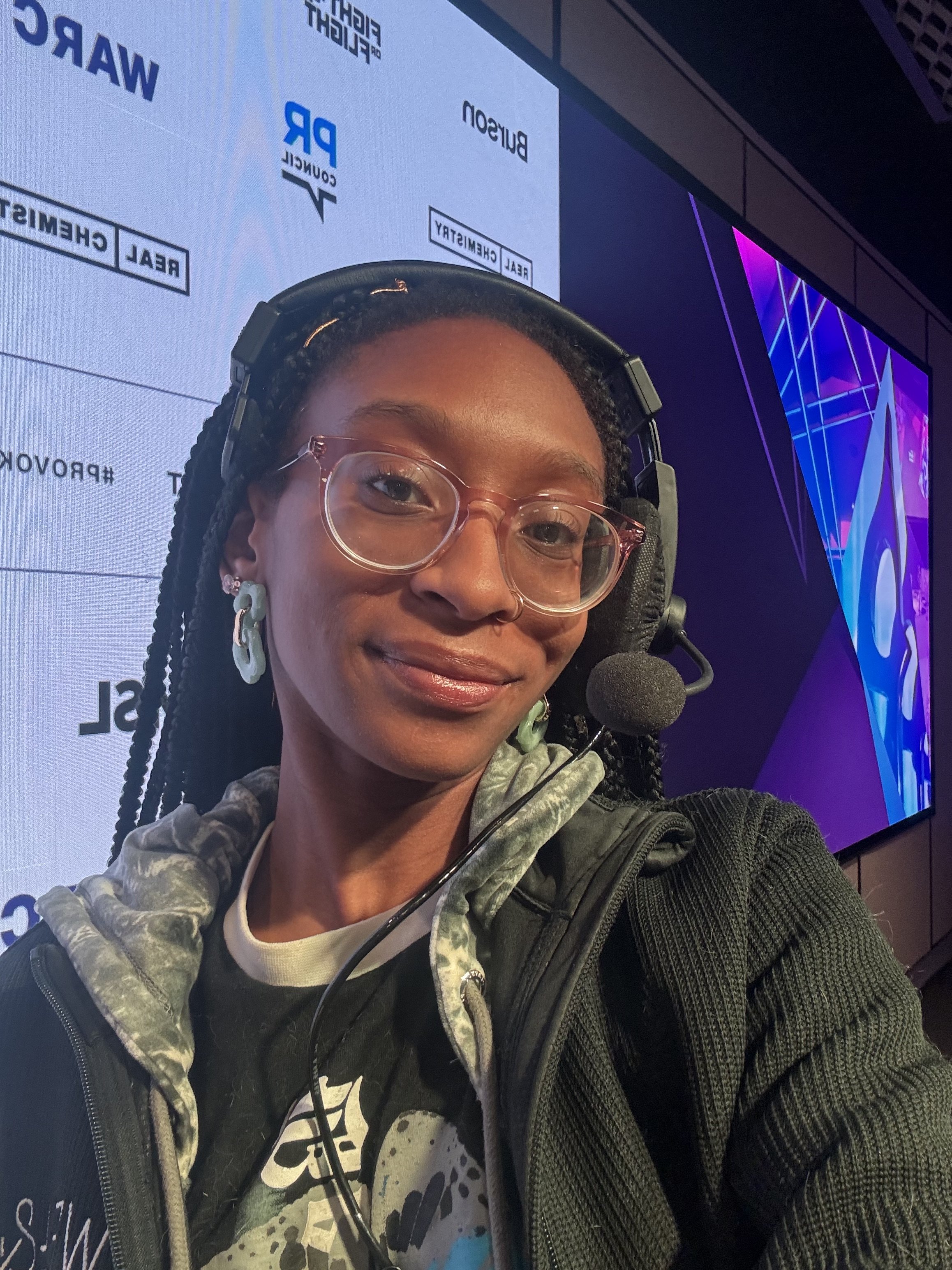 A woman with glasses and braided hair wearing a headset and a jacket, standing in front of a digital screen displaying logos and text related to real estate and a construction company.