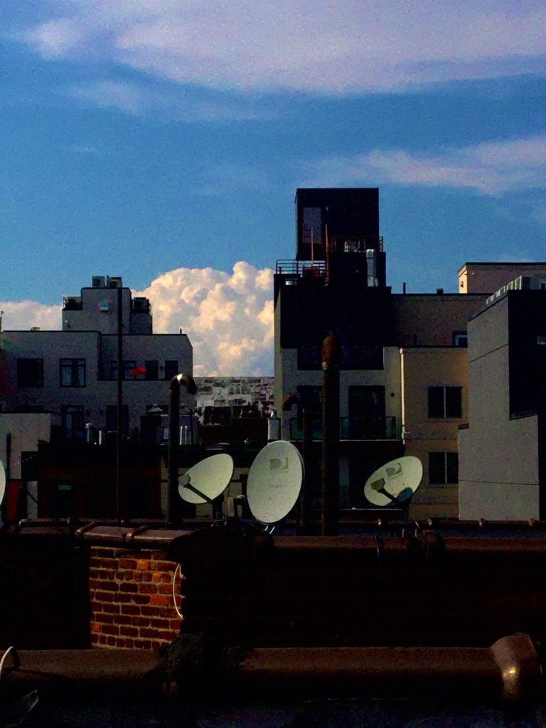 Cityscape with satellite dishes on rooftops, apartment buildings, and a partly cloudy sky during dusk or dawn.