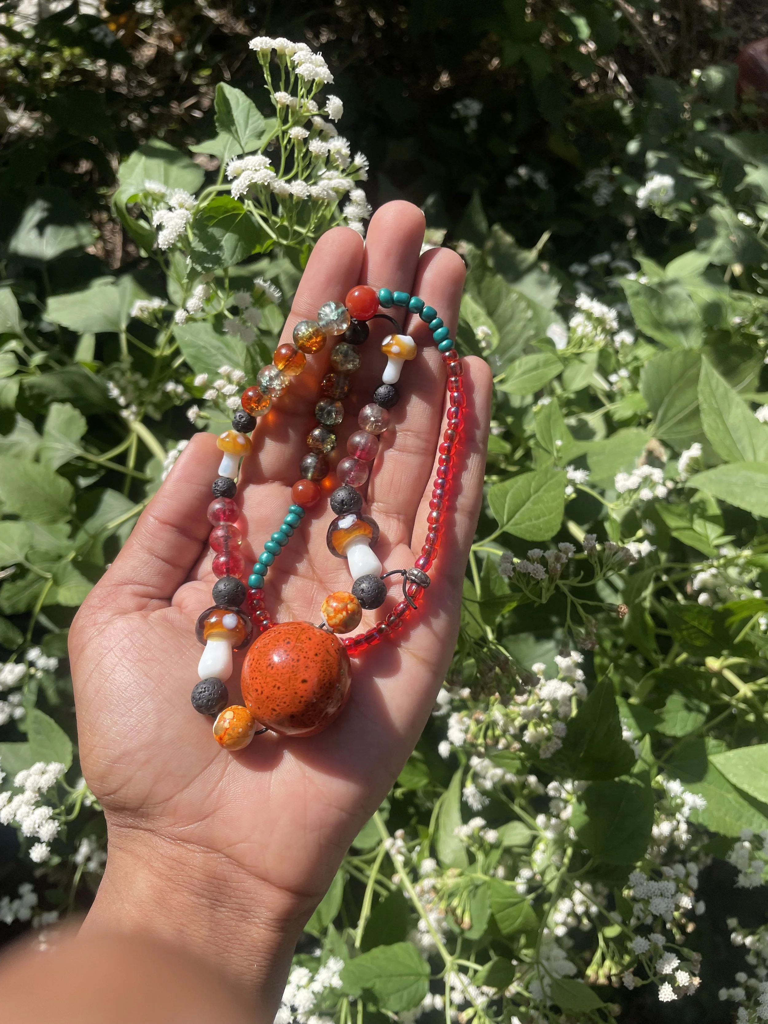 A person holding colorful bead necklaces with a small mushroom pendant in front of green leafy plants and white flowers.