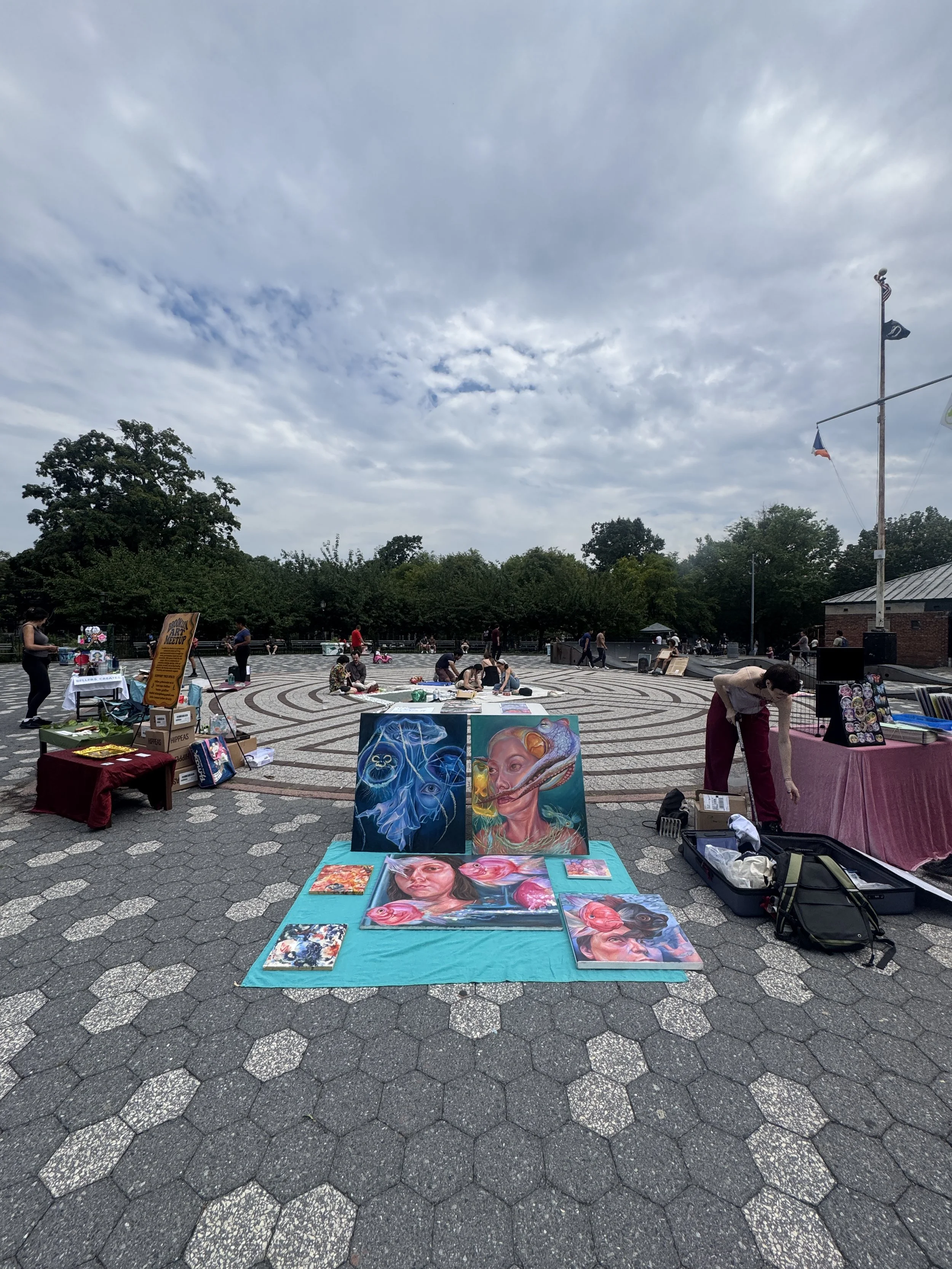 Outdoor art fair with paintings and jewelry booths, people browsing, large trees, cloudy sky, and a flagpole in the background.