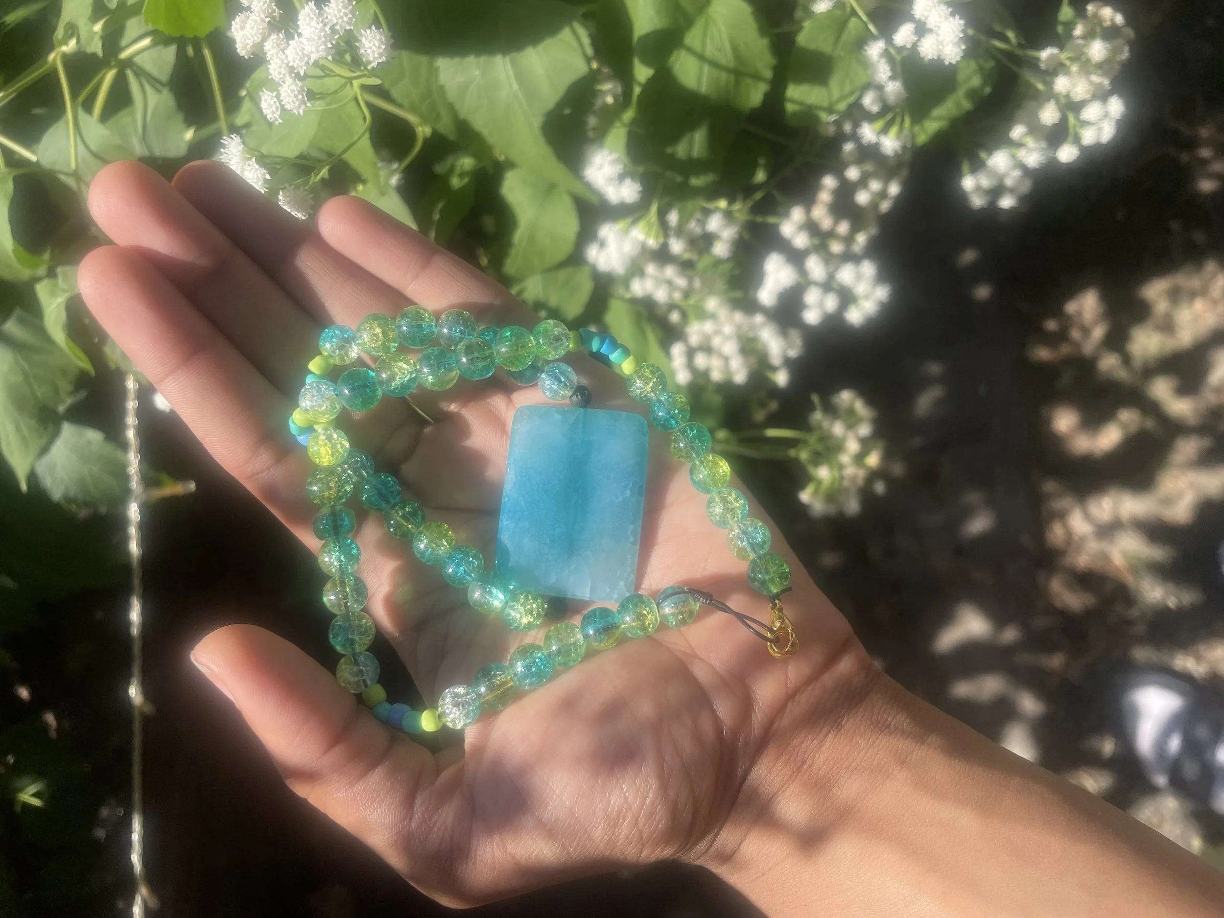 A person's hand holding a blue beaded necklace with a large blue rectangular pendant. The background is filled with green leaves and white flowers, with sunlight illuminating the scene.