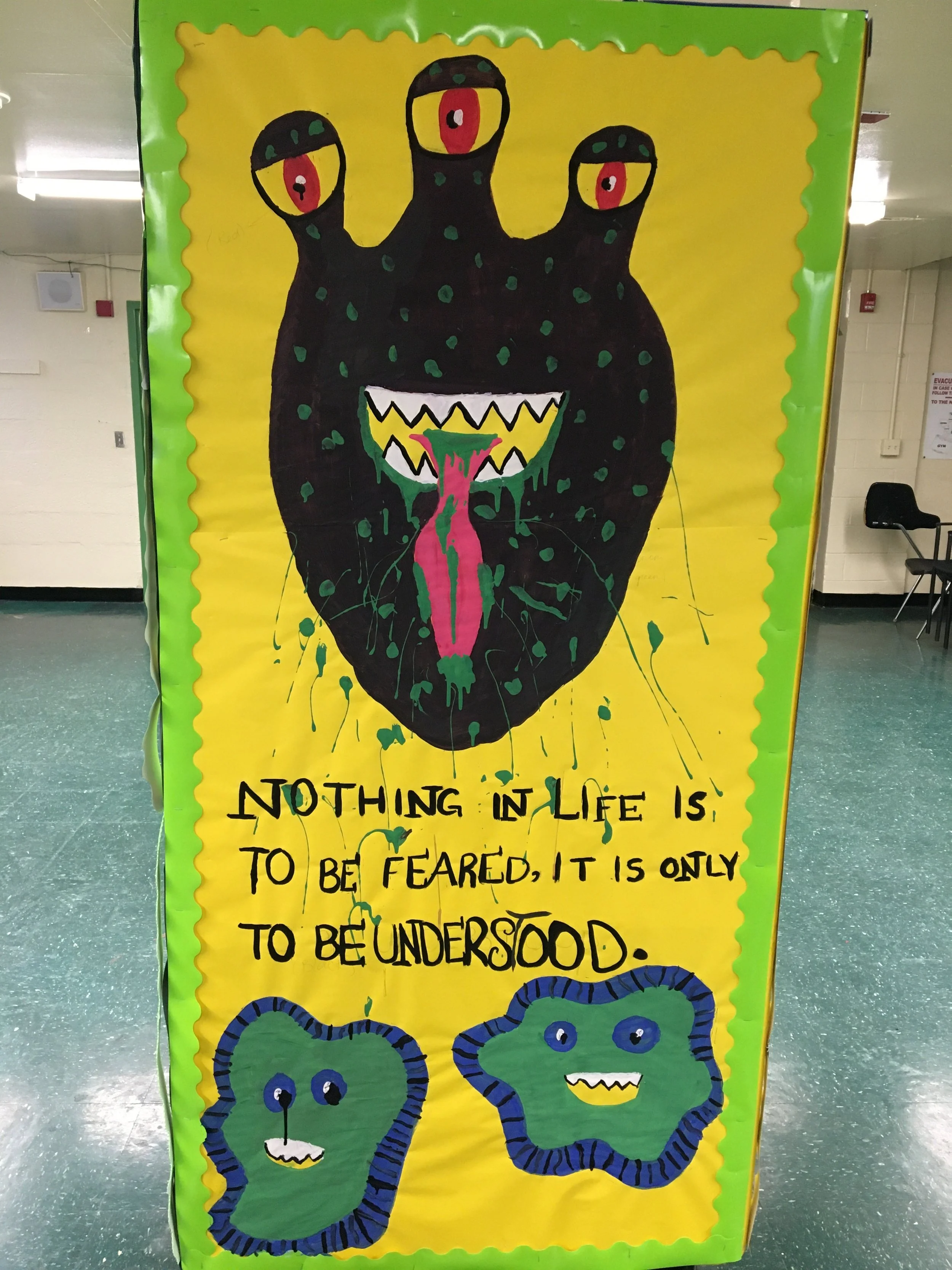 A colorful bulletin board features a large black creature with three eyes, a wide mouth with jagged teeth, and green spots. Its tongue is red and dripping. Below the creature are two cartoonish blue snakes with smiling faces, and a quote that reads: 