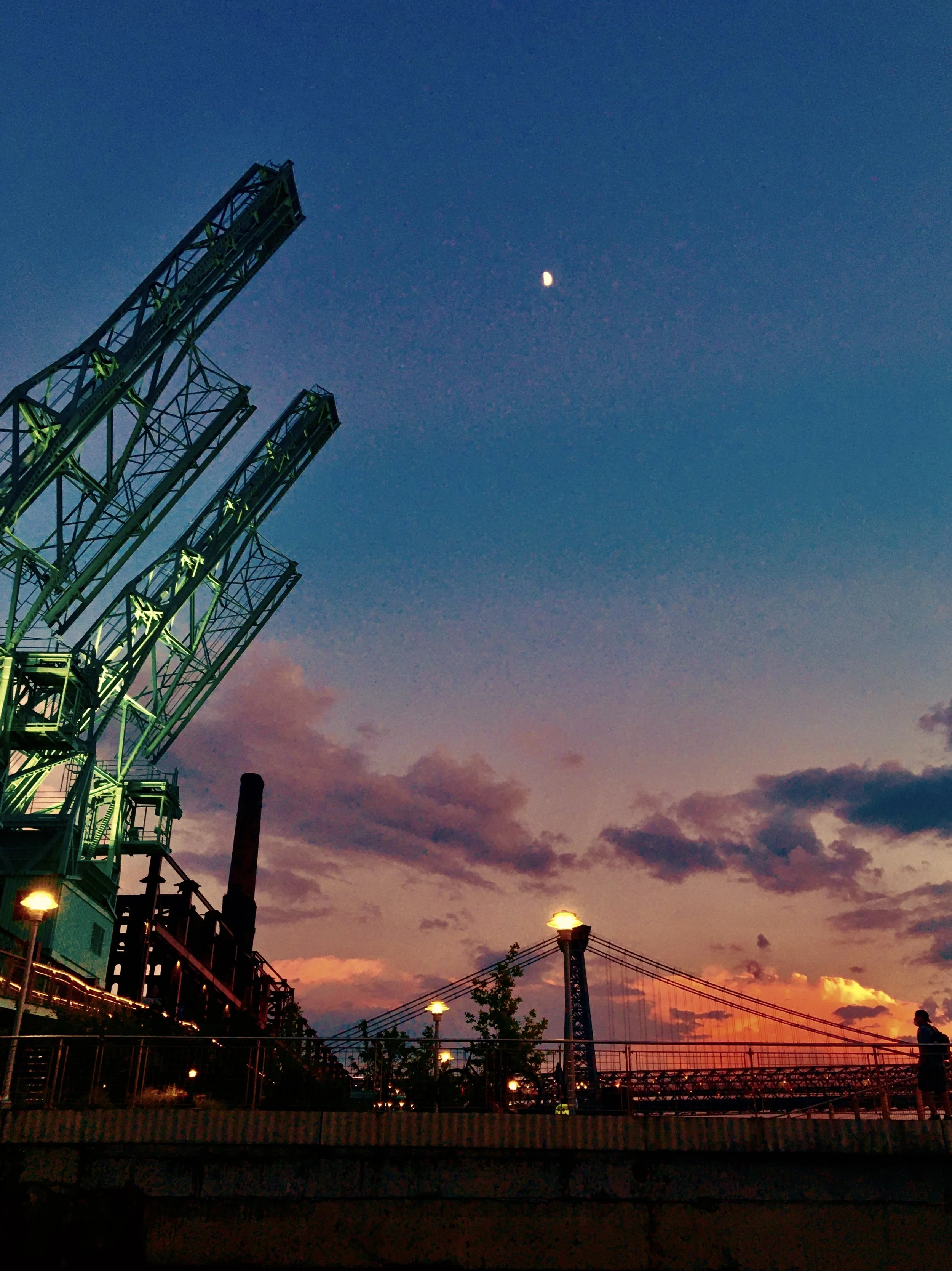 A sky during sunset or dusk with a crescent moon visible. On the left, there is a tall metal industrial structure illuminated with green lights. In the distance, there are a few street lamps and the bridge's towers with cables, as well as some trees and other structures.