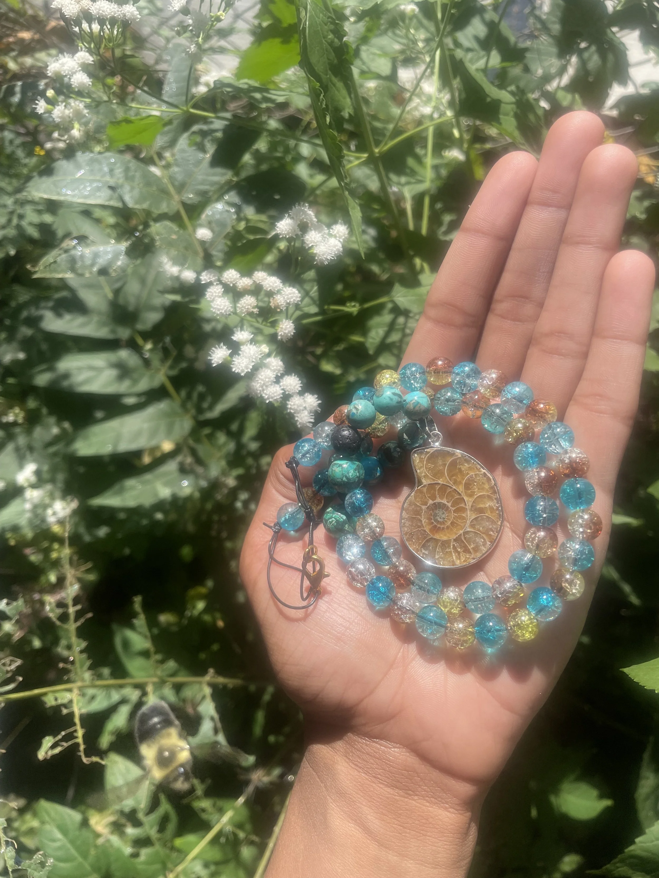 A hand holding colorful beaded jewelry and a shell in front of green leafy plants and white flowering plants.