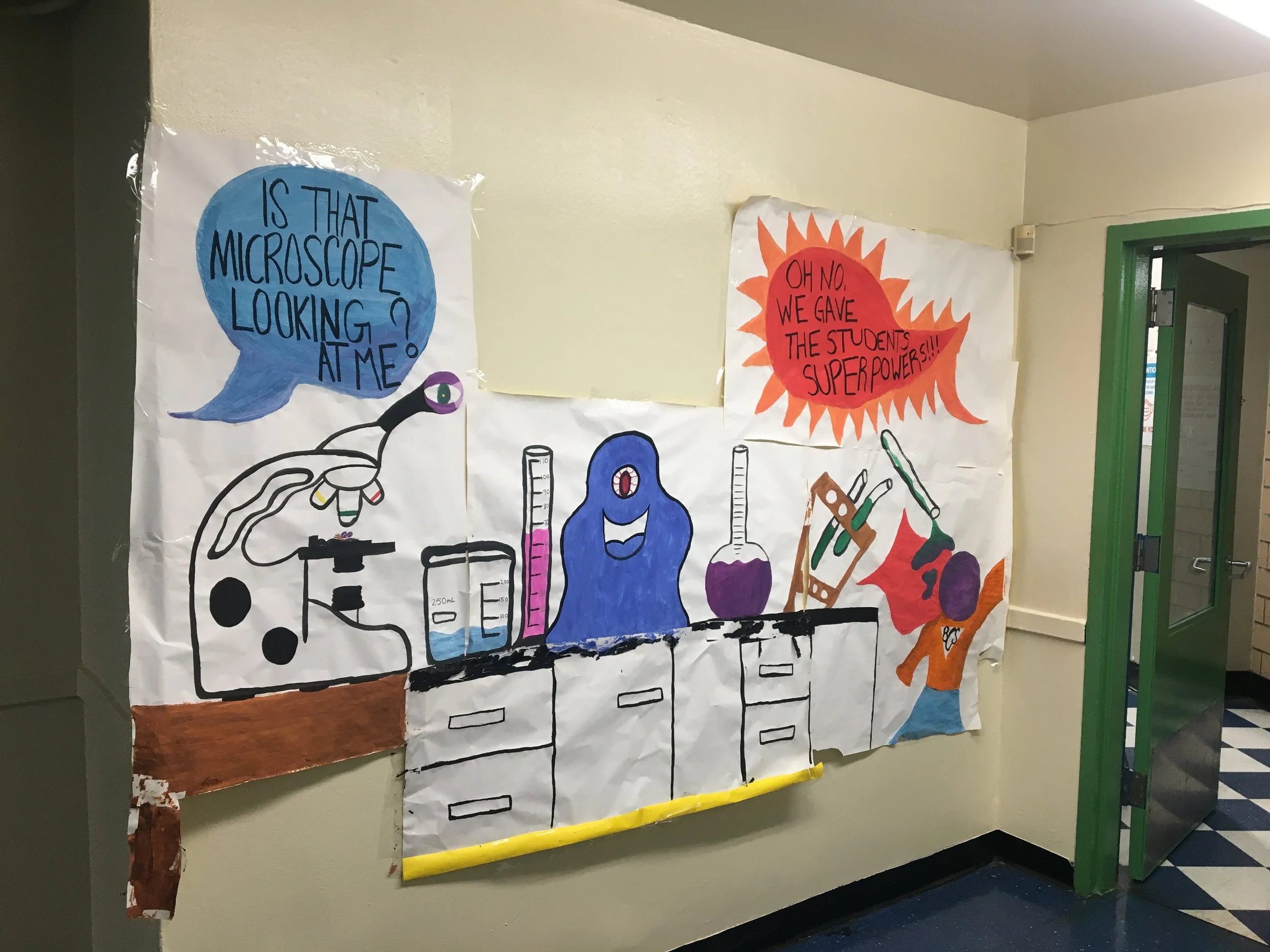 Colorful poster on a school hallway wall features science-related illustrations. The left side shows a blue speech bubble with the text 'Is that microscope looking at me?' and a drawing of a microscope. The right side has a red speech bubble with the