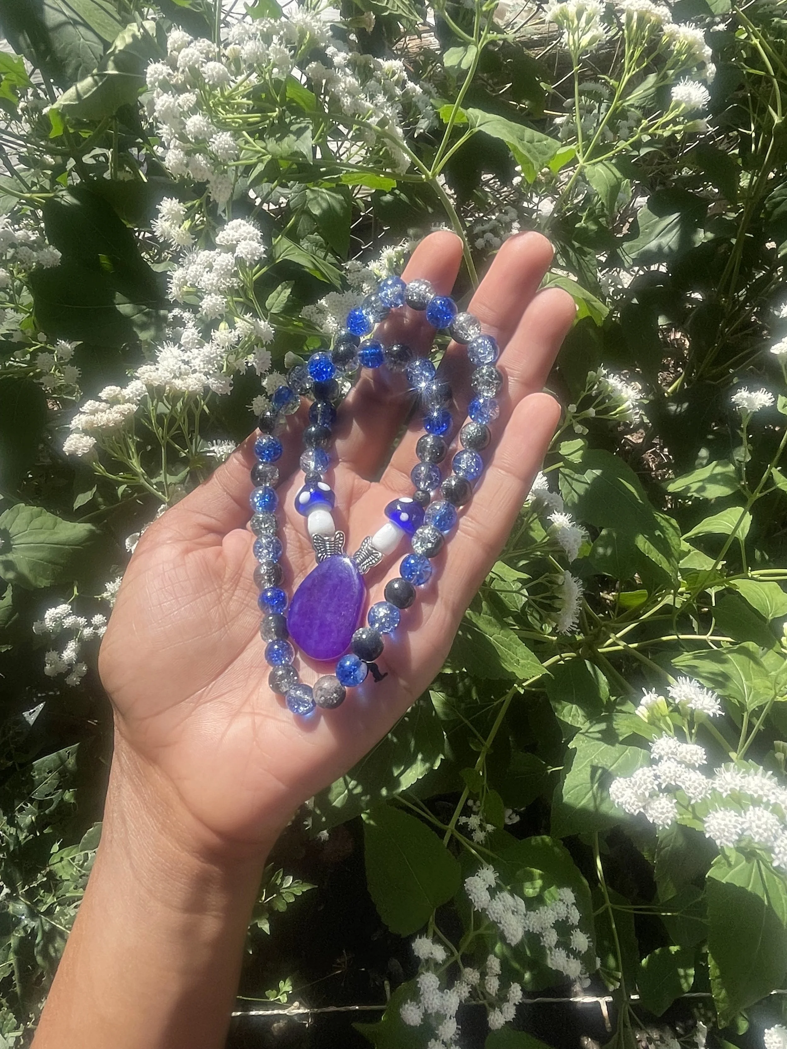 A hand holding a blue and gray beaded bracelet with a purple stone and butterfly charm in front of white flowering plants.