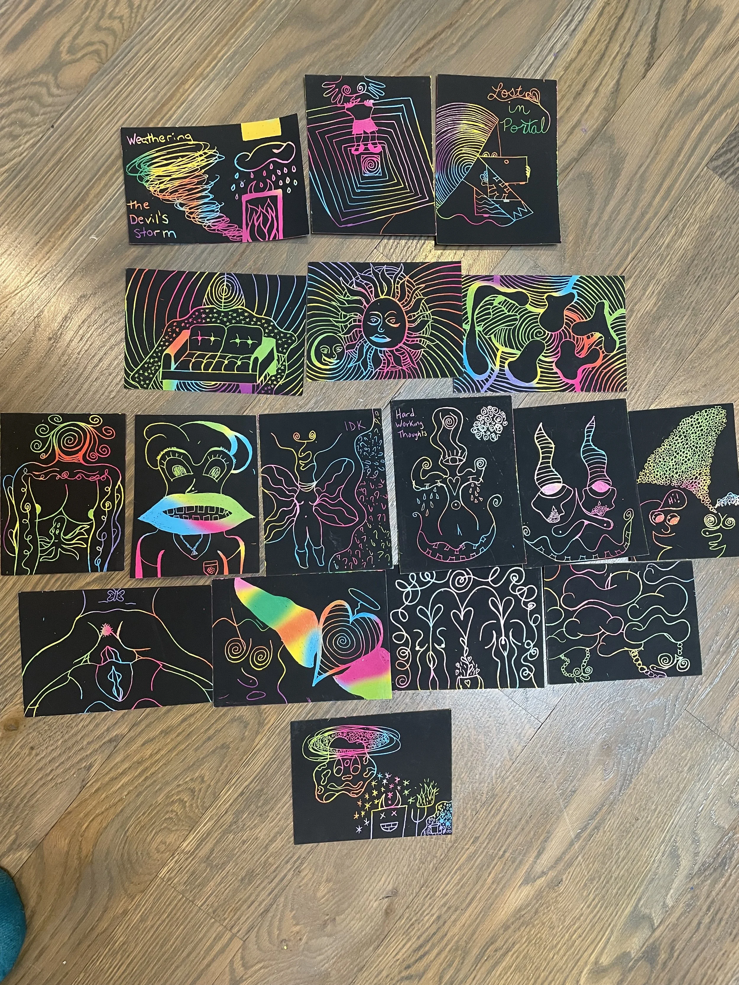 A collection of colorful, neon-style illustrations on black background cards, depicting various whimsical characters, abstract patterns, and scenes, arranged in rows on a wooden floor.