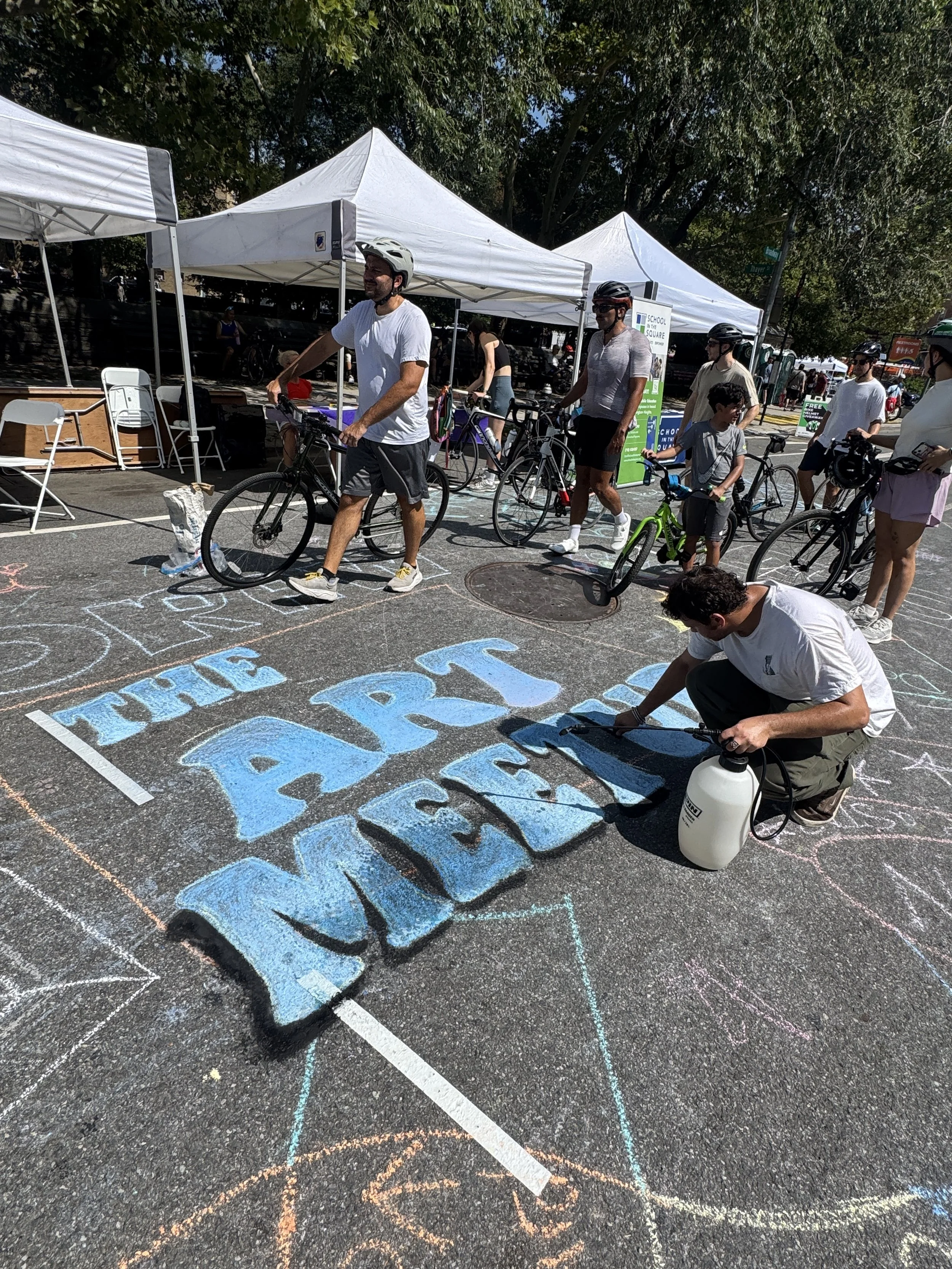 People participating in an outdoor chalk art event on a parking lot, with tents, drawings, and bicycles. A man is kneeling and working on chalk art on the pavement.