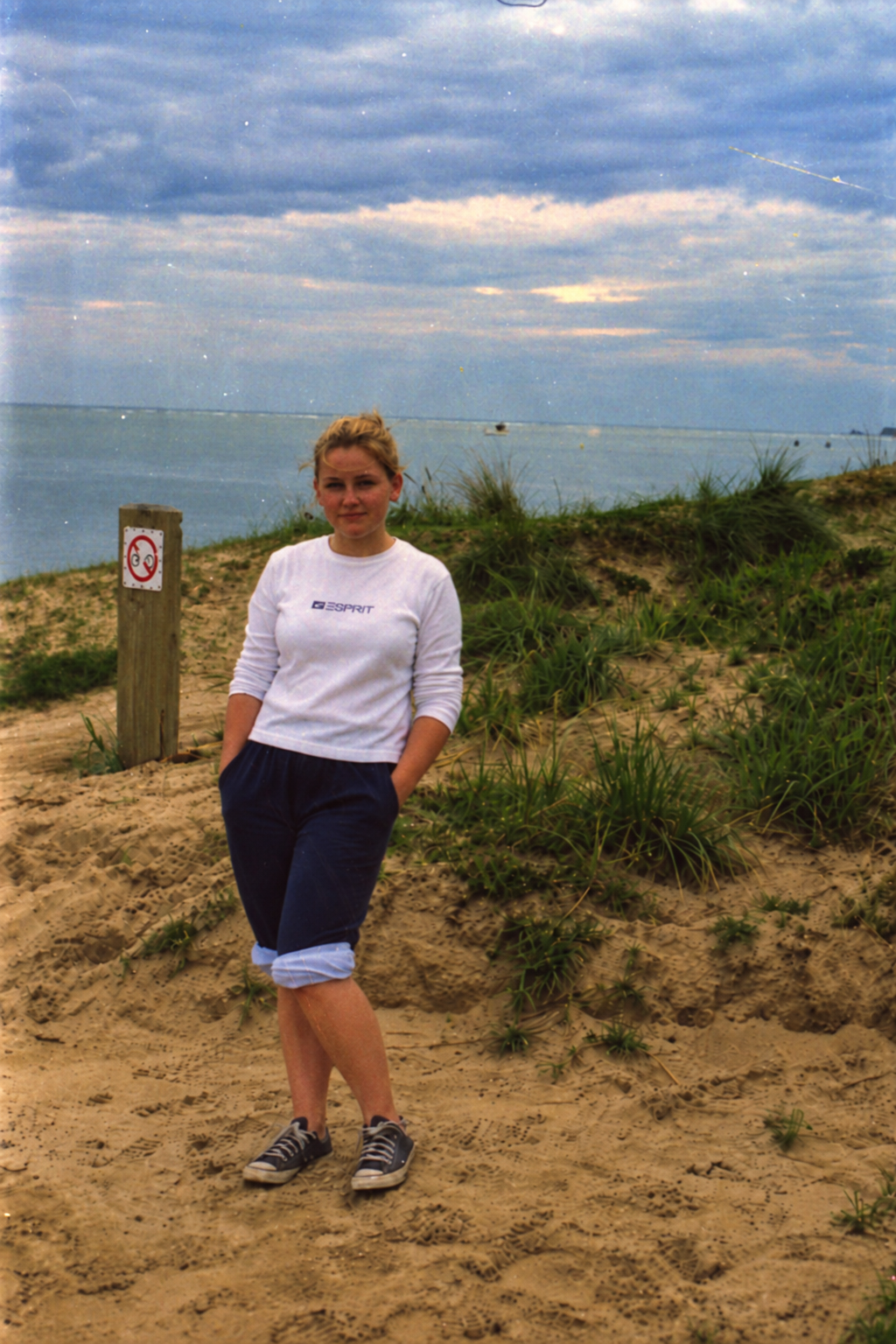 A young woman standing on a sandy beach near dunes and grass, with the ocean and cloudy sky in the background.