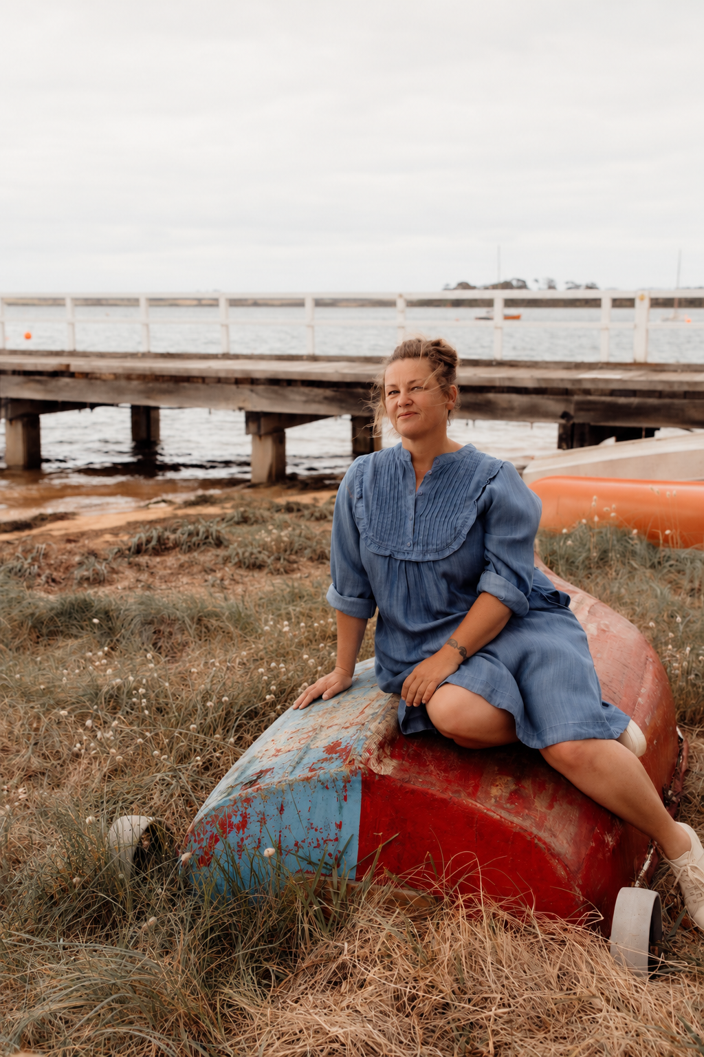 A woman in a blue dress sitting on an old red and blue boat on a grassy area near a body of water, with a wooden pier and cloudy sky in the background.