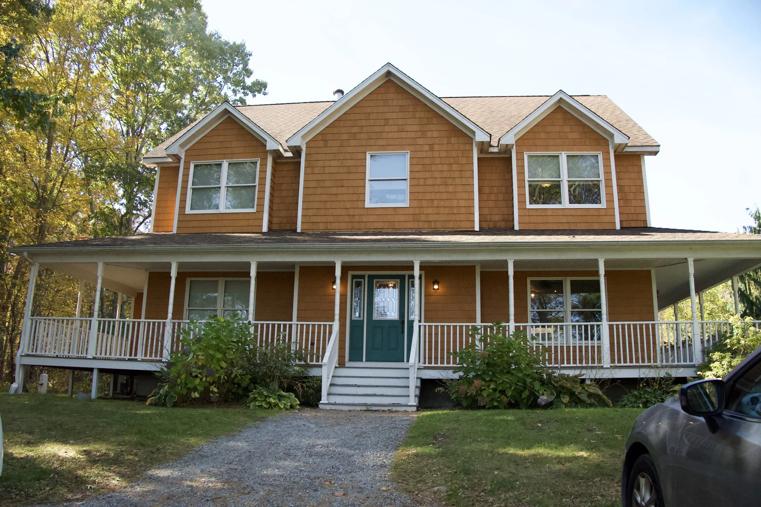 A two-story house with orange siding, white trim, and a green front door, featuring a front porch with white railings and steps, surrounded by trees and a gravel driveway.