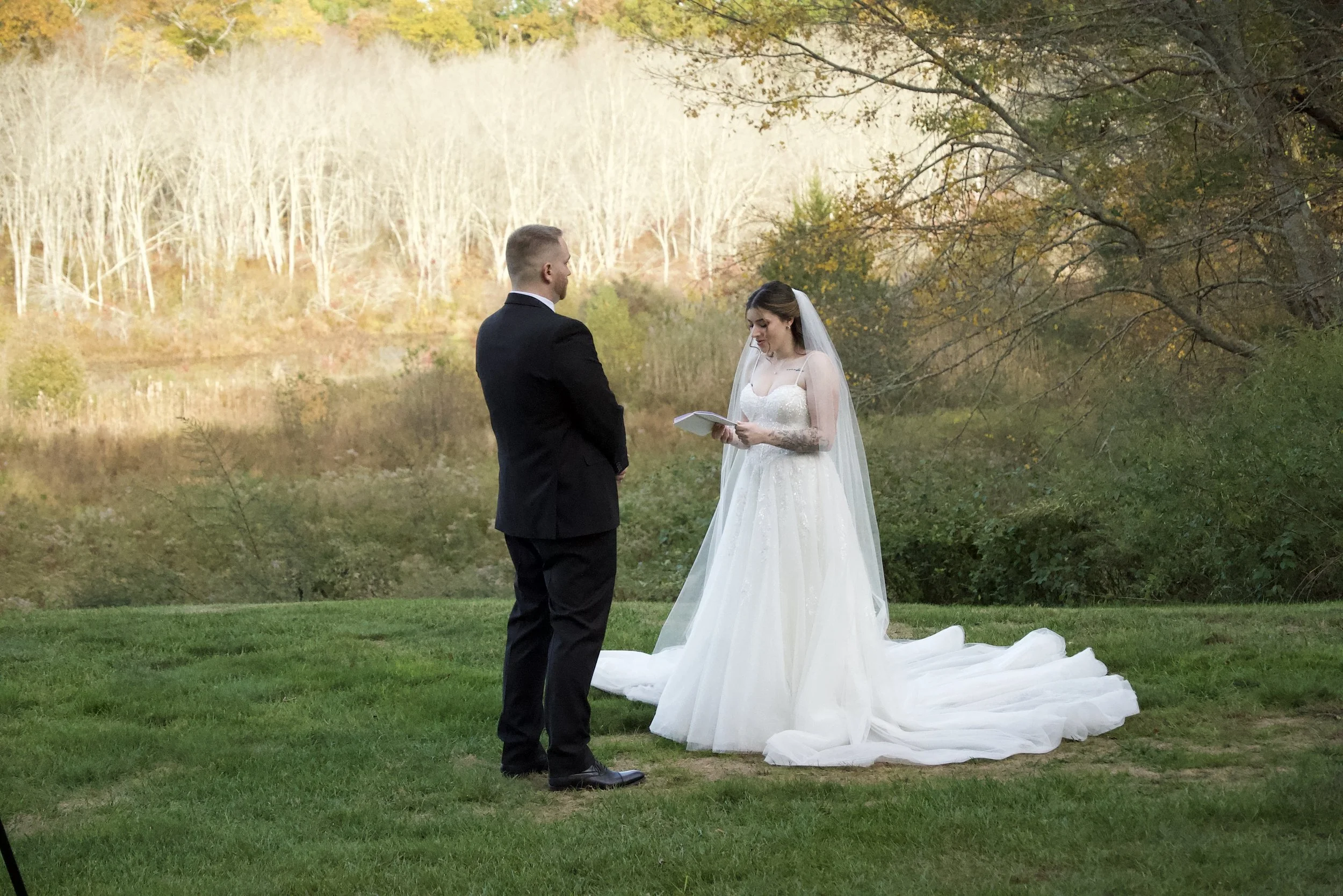 A couple standing outdoors during a wedding ceremony by a lake surrounded by fall trees. The bride is reading vows, wearing a white wedding dress with a long train and veil, while the groom is in a black suit.