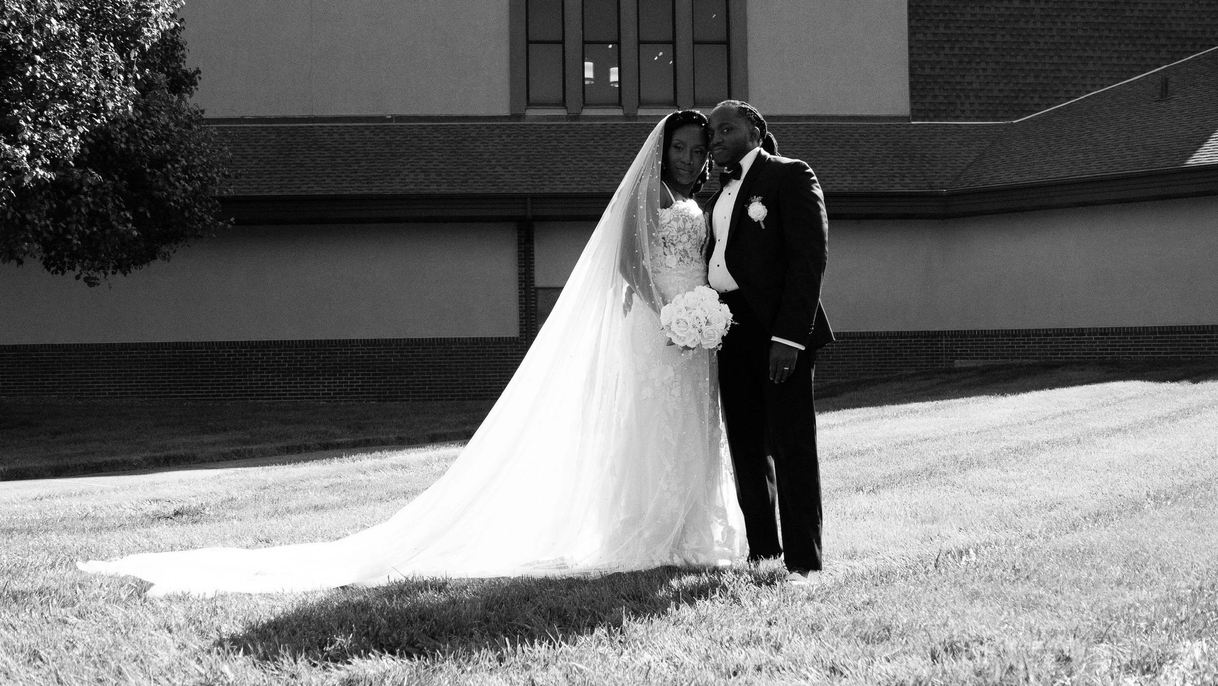 Black and white photo of a bride and groom standing together outdoors, dressed in wedding attire, with a building and tree in the background.