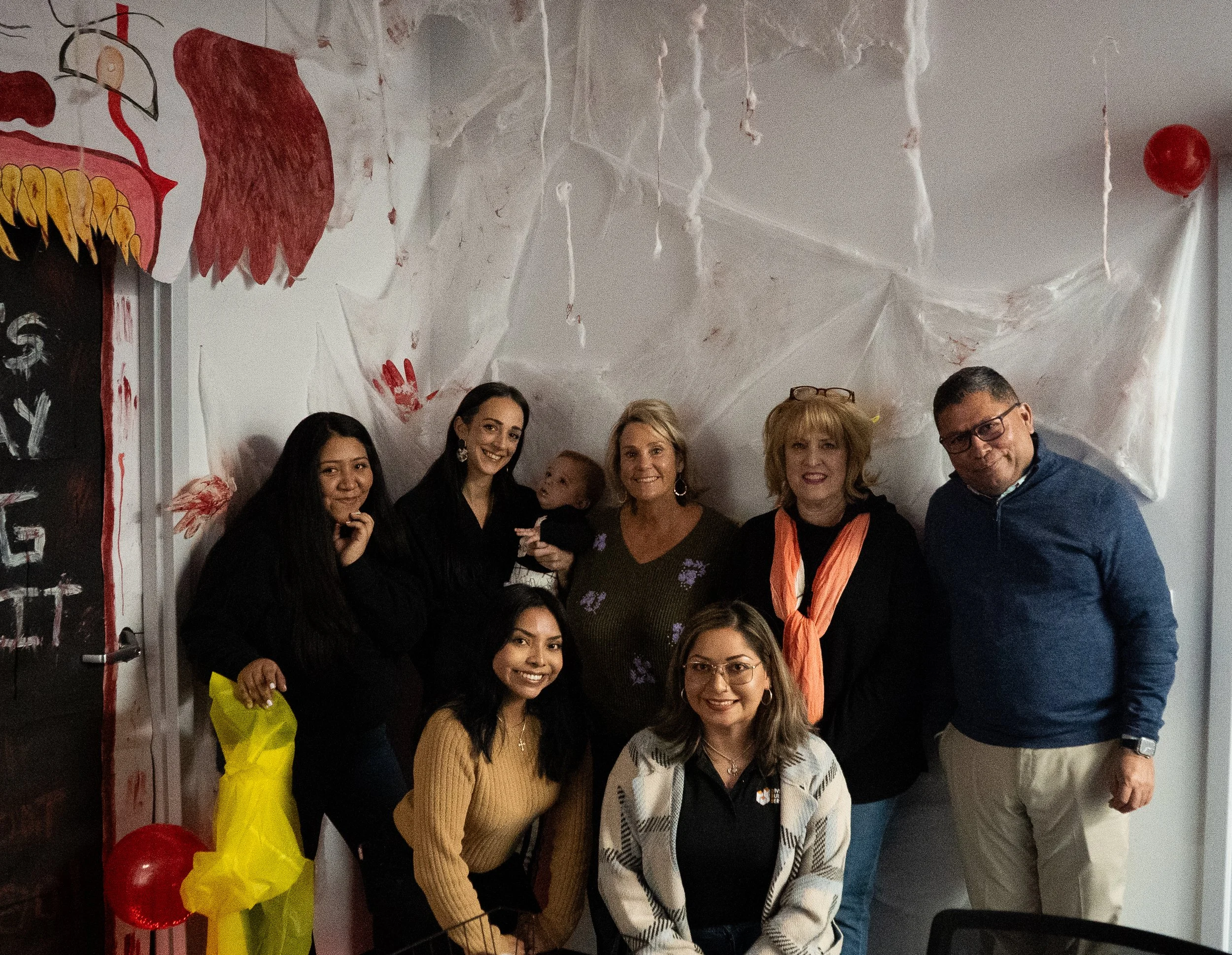 A group of ten people posing together indoors at a celebration or gathering, smiling, with decorations including red and yellow balloons and a large spider web decoration on the ceiling.
