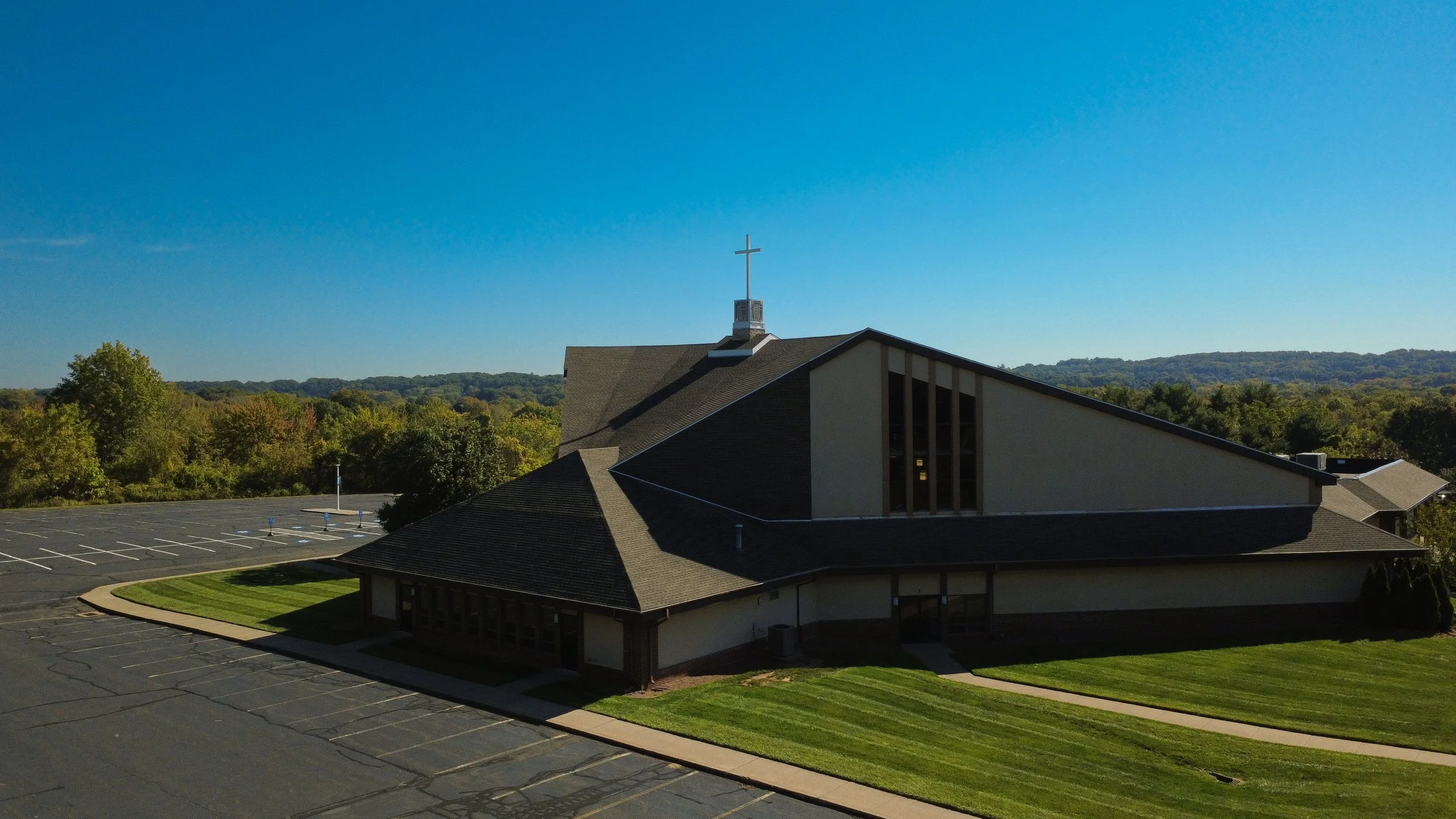 A church with a cross on top, surrounded by trees and an empty parking lot under a clear blue sky.