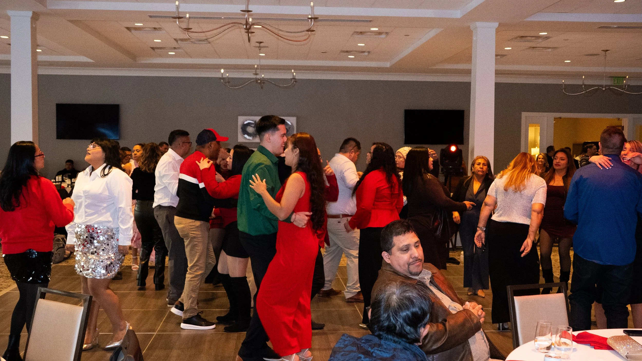 People dancing and socializing at a party in a banquet hall decorated in red, green, and black outfits, with some wearing festive or formal attire.