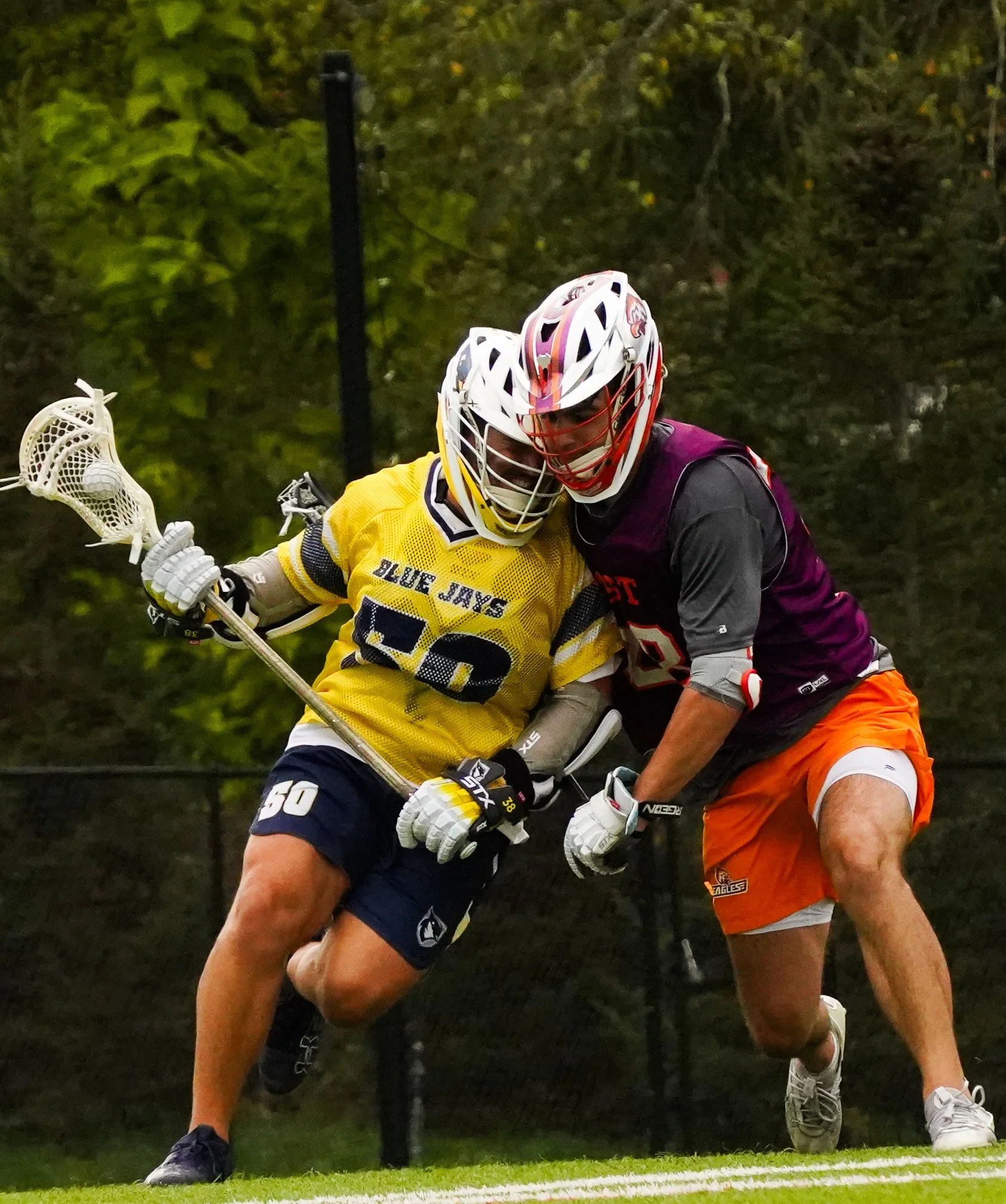 Two lacrosse players in a close game, one wearing a yellow jersey and the other in a dark jersey, both wearing helmets, gloves, and athletic gear, actively competing near the ground on a grass field with a blurred background of trees.