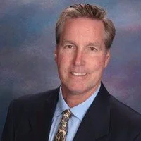 Portrait of a middle-aged man with gray hair, wearing a business suit and tie, smiling against a blurred, colorful background.