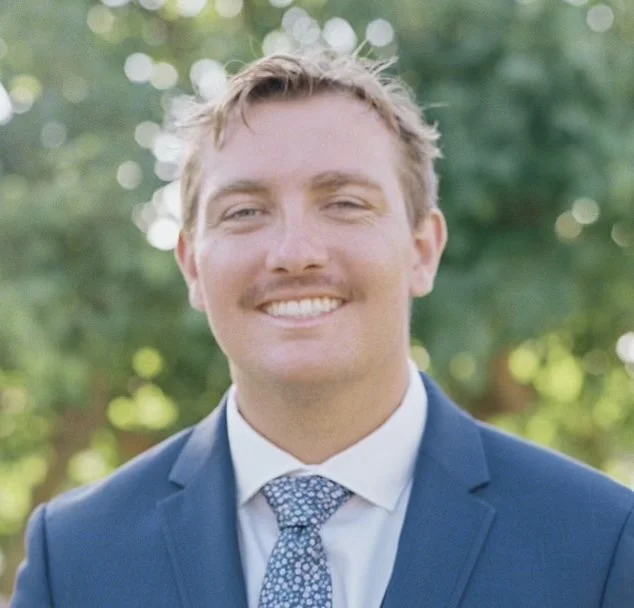 A young man in a blue suit and white shirt smiling outdoors with blurred trees and sunlight in the background.