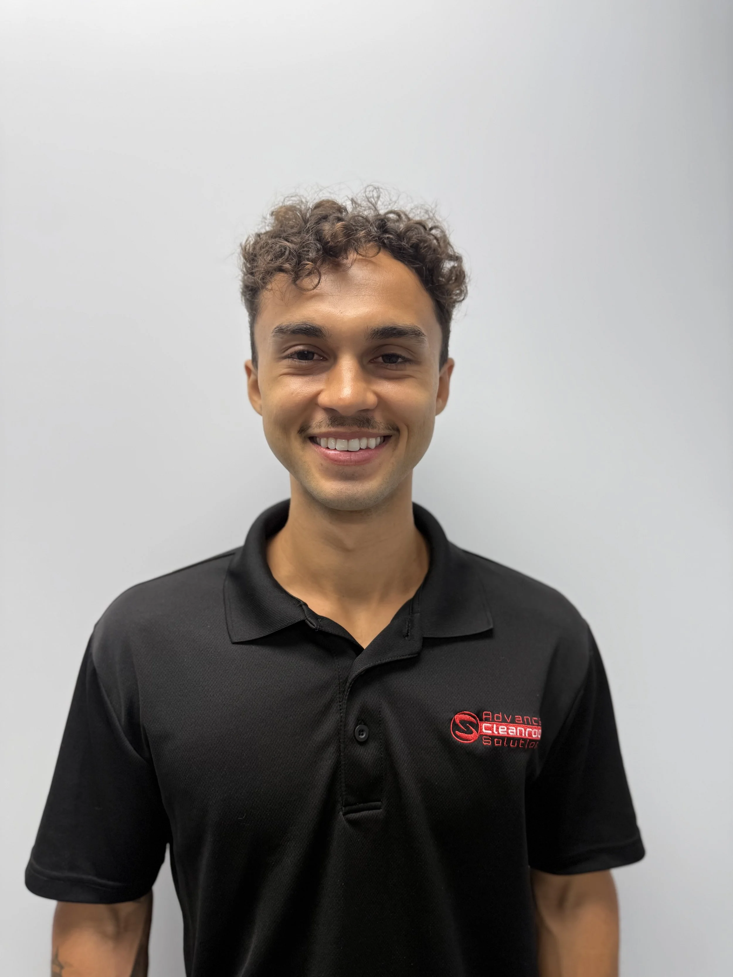 A young man with curly brown hair, wearing a black polo shirt with the logo 'Advance Cleaning Solution' on the chest, smiling against a plain white wall background.