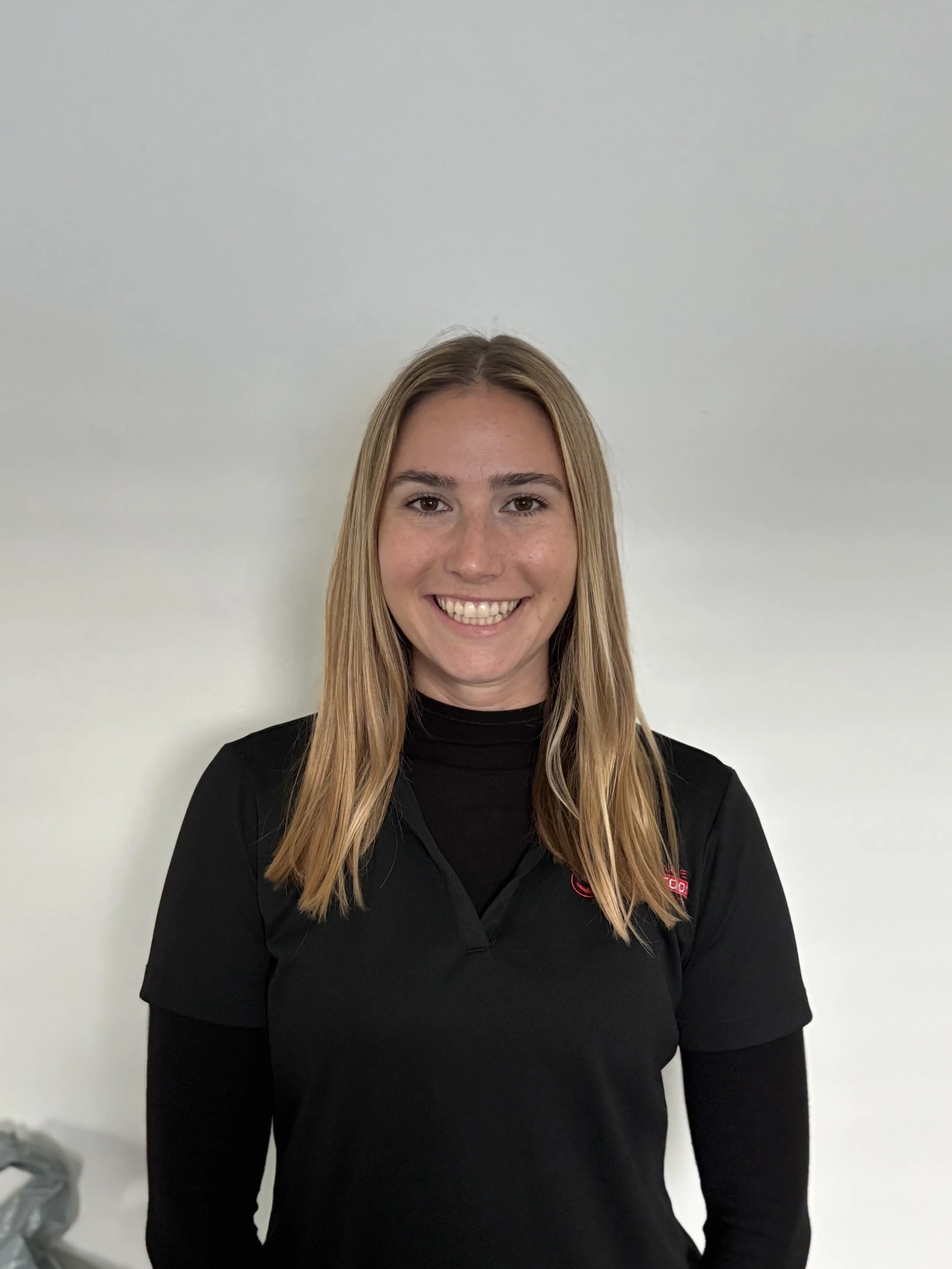 Young woman with long blonde hair smiling, wearing a black shirt with a logo on the left chest, standing against a plain white wall.
