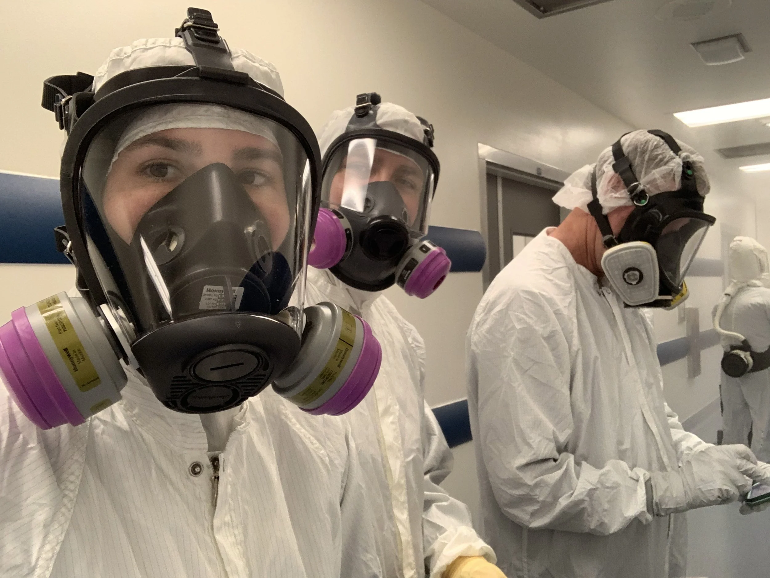 Three healthcare workers wearing full protective suits, masks, and head coverings in a clinical setting.