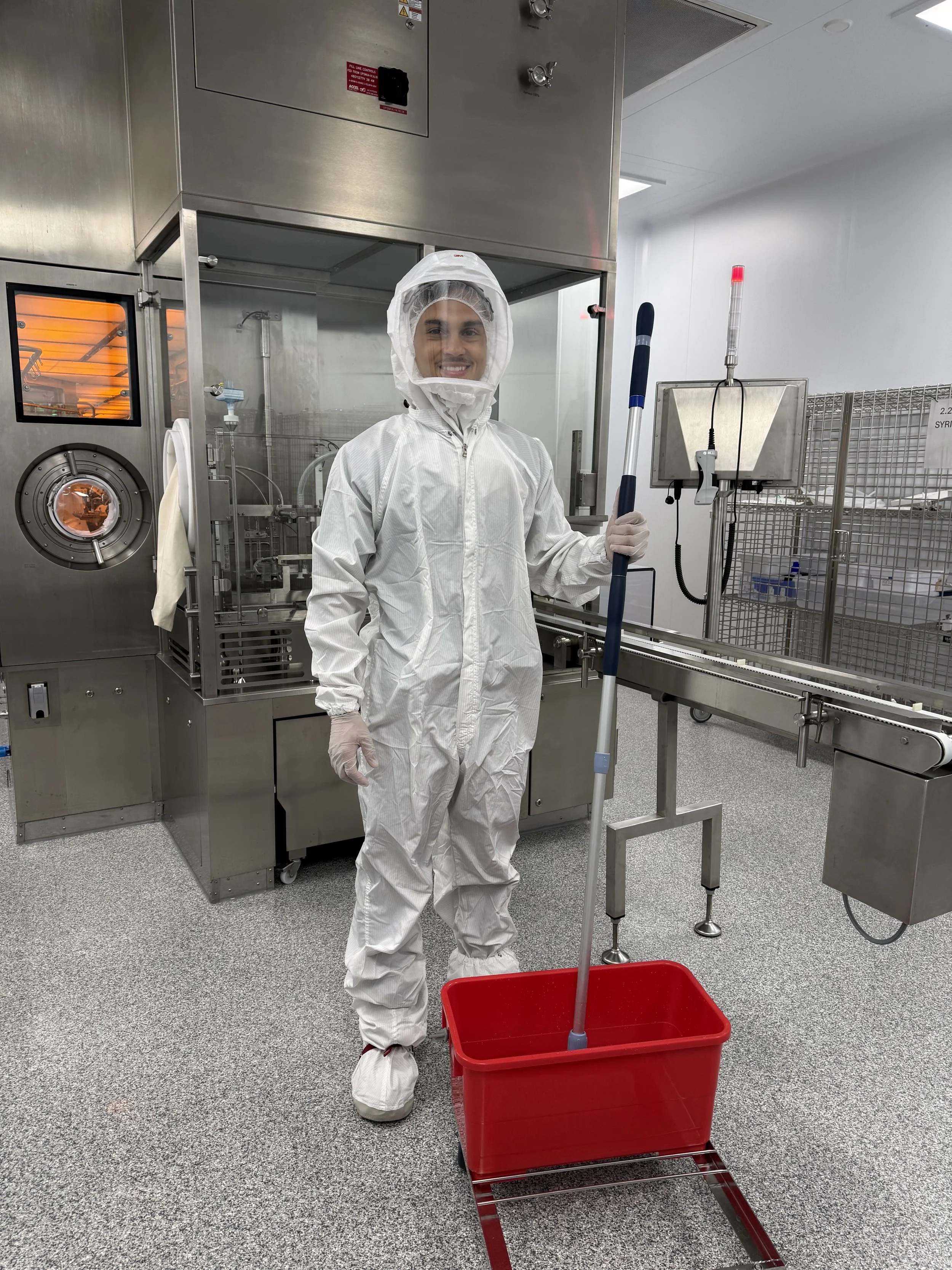 Person in protective clothing standing in a laboratory or cleanroom, holding a mop and bucket for cleaning. The person is smiling and wearing a full-body white protective suit, gloves, and a head covering. There is industrial equipment and a metal cart in the background.