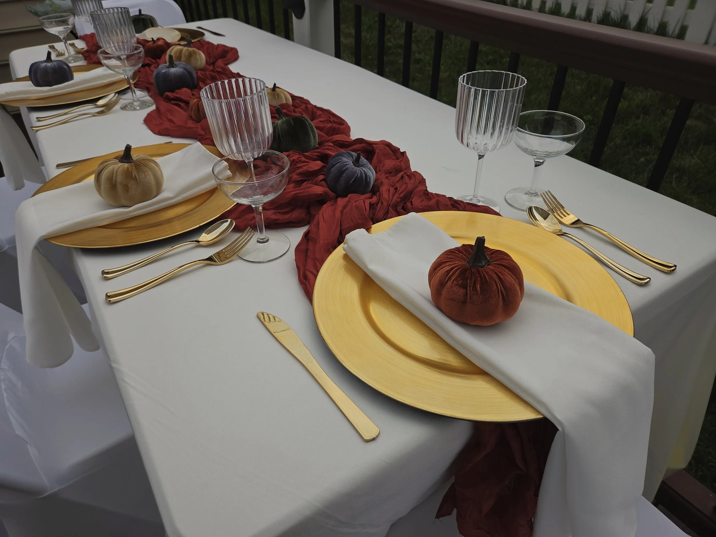 A dining table set for a meal with a white tablecloth, gold flatware, and gold-rimmed glassware. The table features a fall-themed centerpiece with small pumpkins in various colors placed on a red fabric runner. Each place setting has a white napkin on a yellow plate.