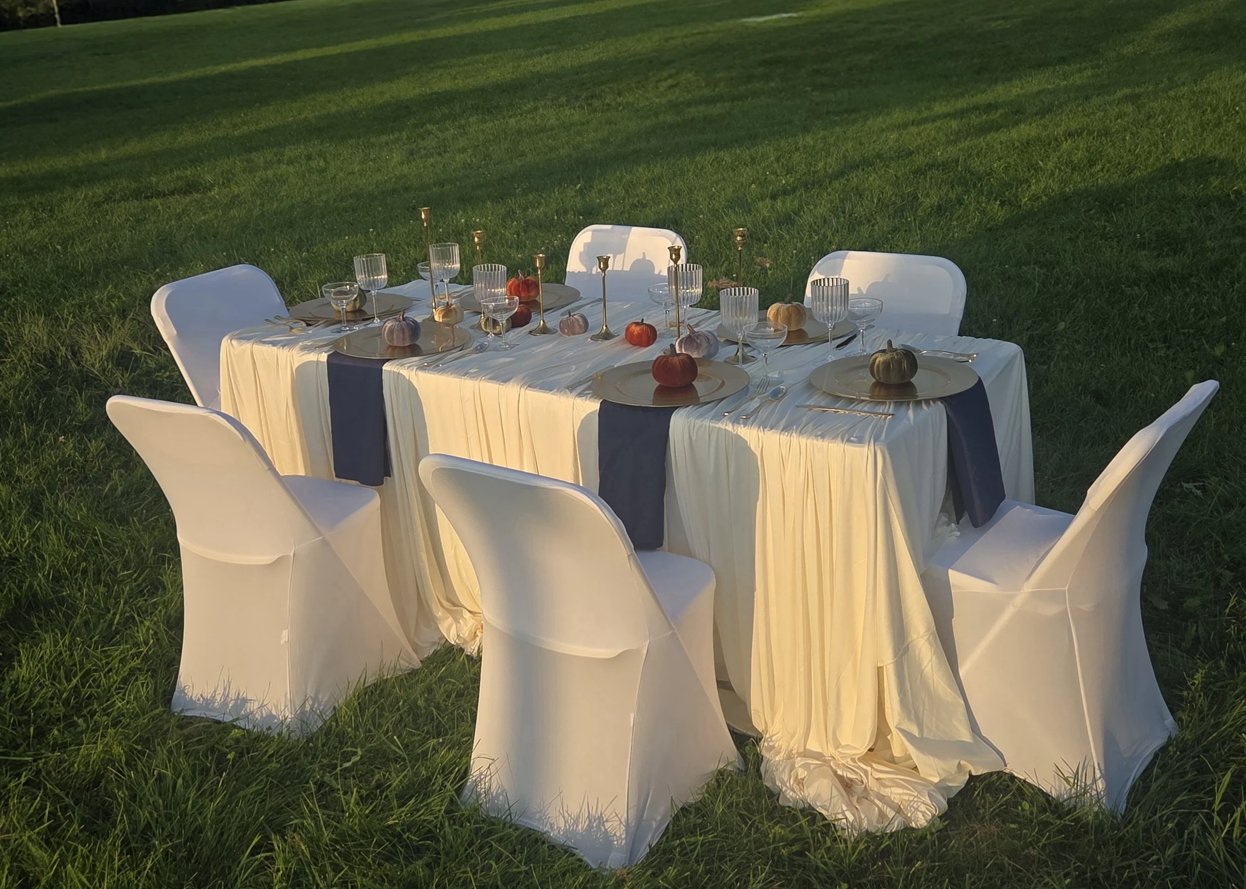 An outdoor dining table set on grass, decorated with small pumpkins, gold candlesticks, and glassware, during late afternoon sunlight.
