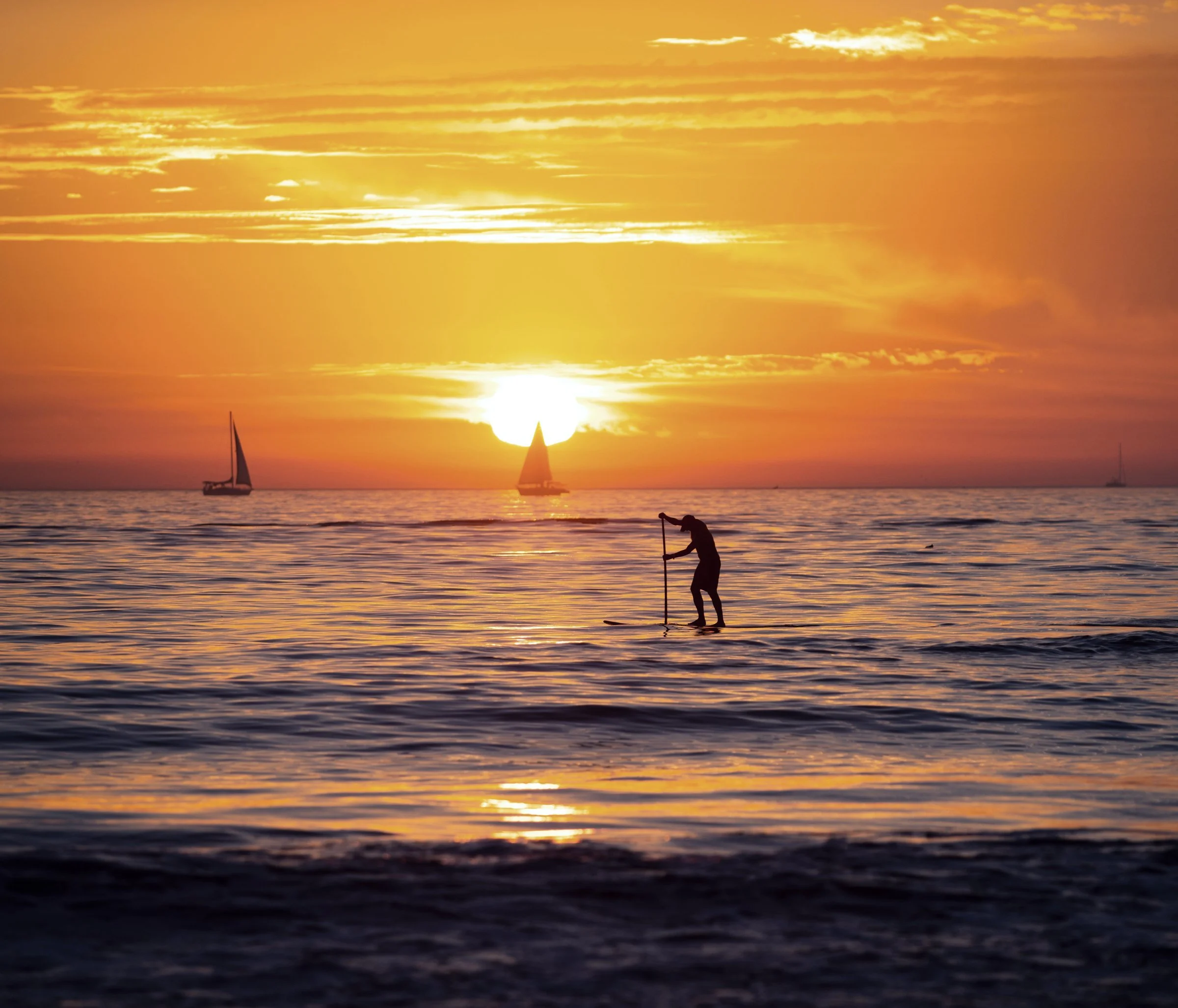 Paddleboarder at sunset on calm Florida water with sailboats on the horizon
