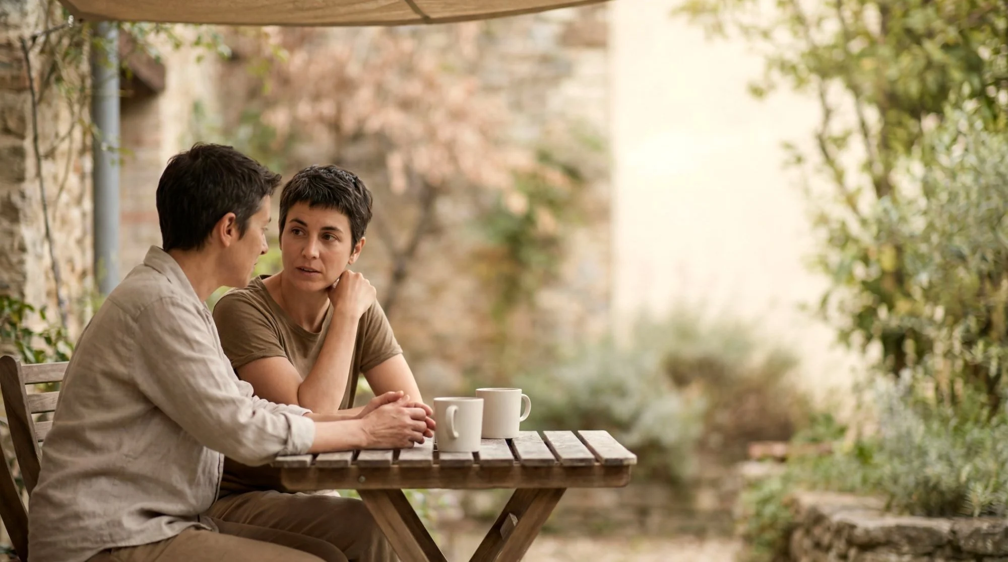 Two women talking over coffee at a wooden table in a quiet garden courtyard