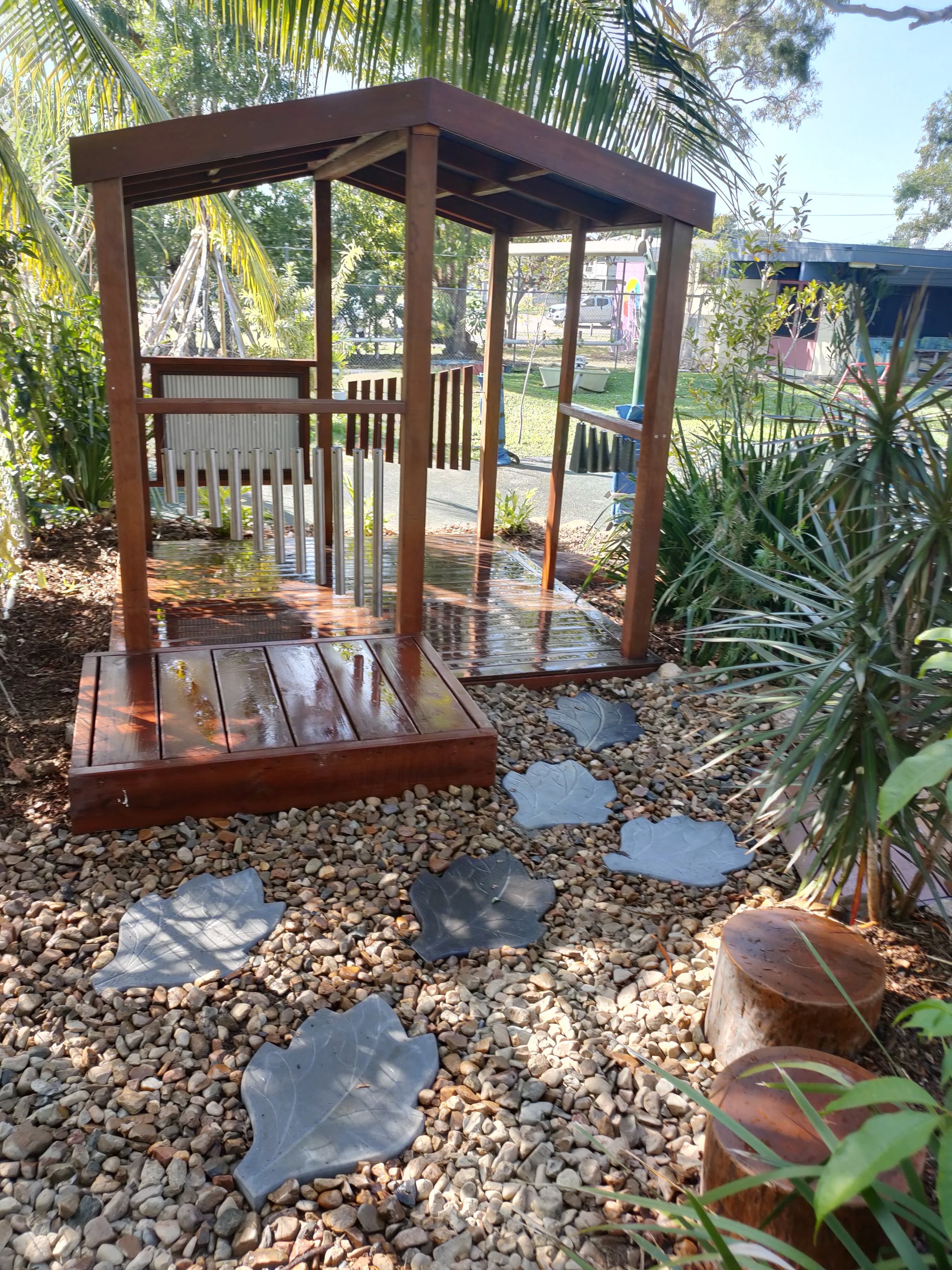 A small wooden playhouse with a peaked roof, located outdoors in a garden surrounded by green plants and decorated with leaf-shaped stepping stones on a gravel pathway.