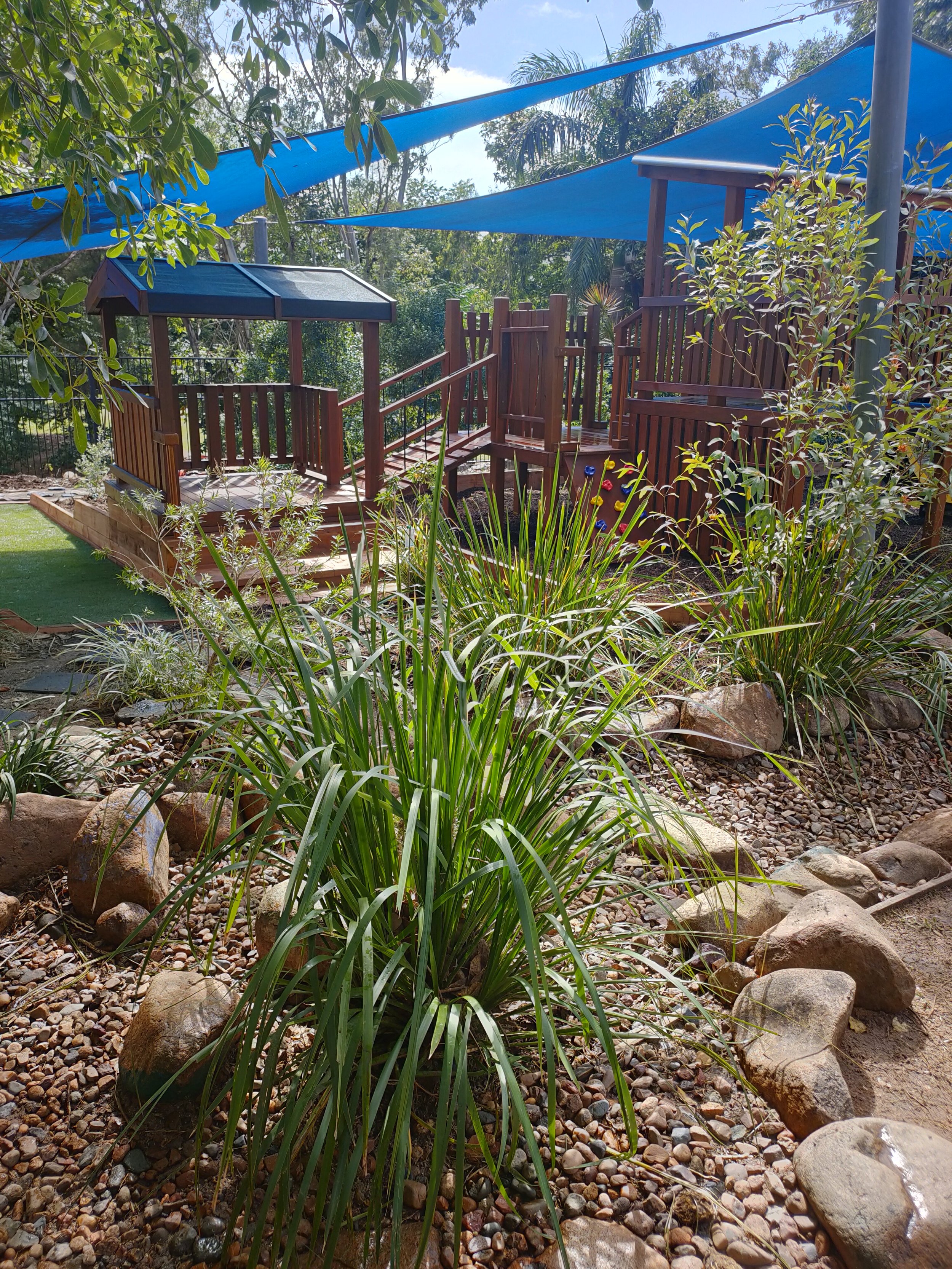 A backyard garden with a wooden playhouse, blue shade sails, and lush green plants, rocks, and gravel on the ground, with trees and a fence in the background.