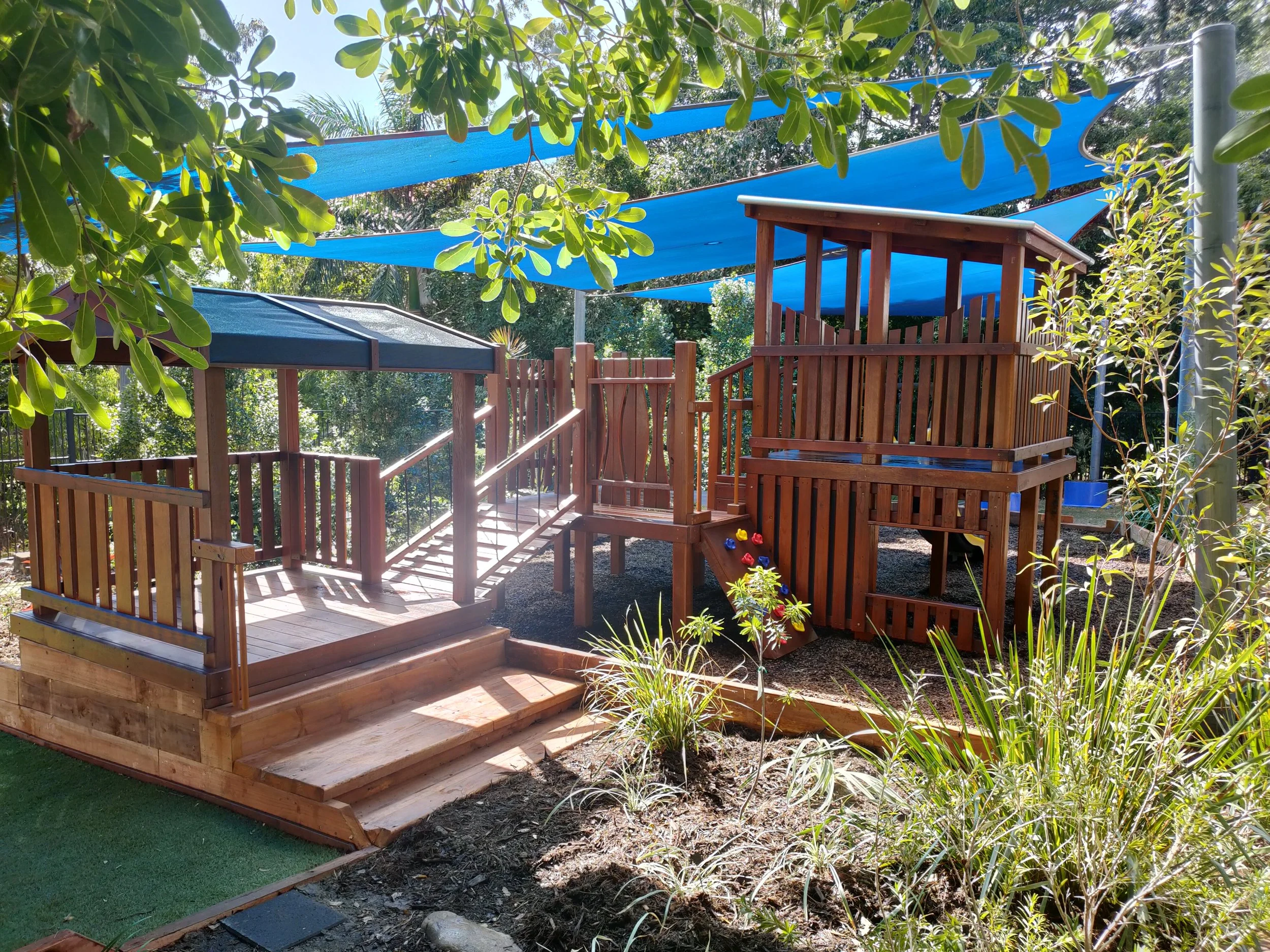 A wooden play structure with stairs and a raised platform in a backyard, shaded by blue sun shades and surrounded by green trees and plants.