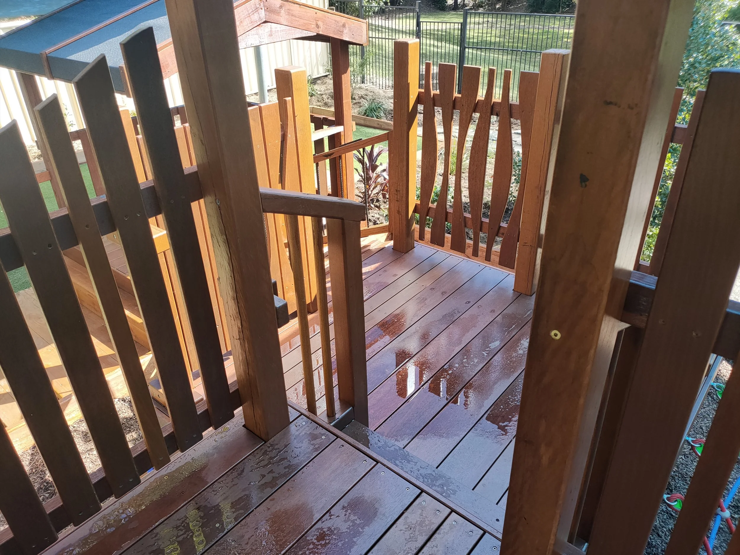View of a wooden deck with a section of fencing and garden in the background, showing part of a playground or backyard area.