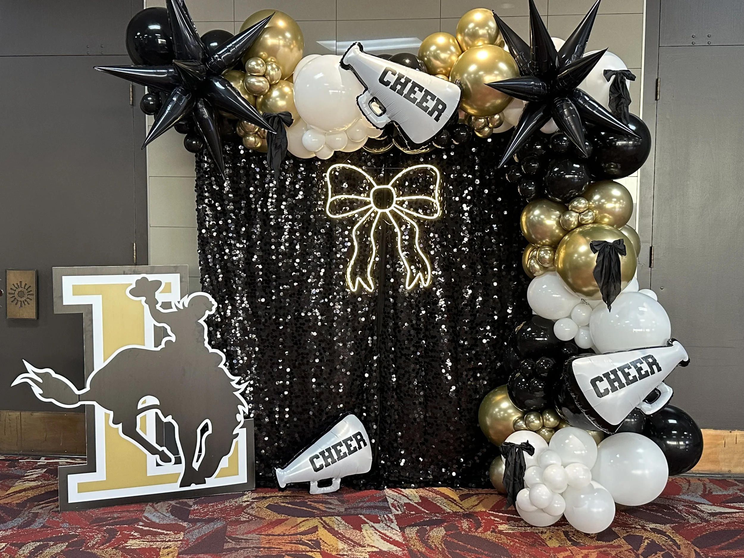 Decorative display with black, white, and gold balloons, some with bows, and banners with cheerleading themes, including a neon outline of a cheer bow and a silhouette of a cheerleader.