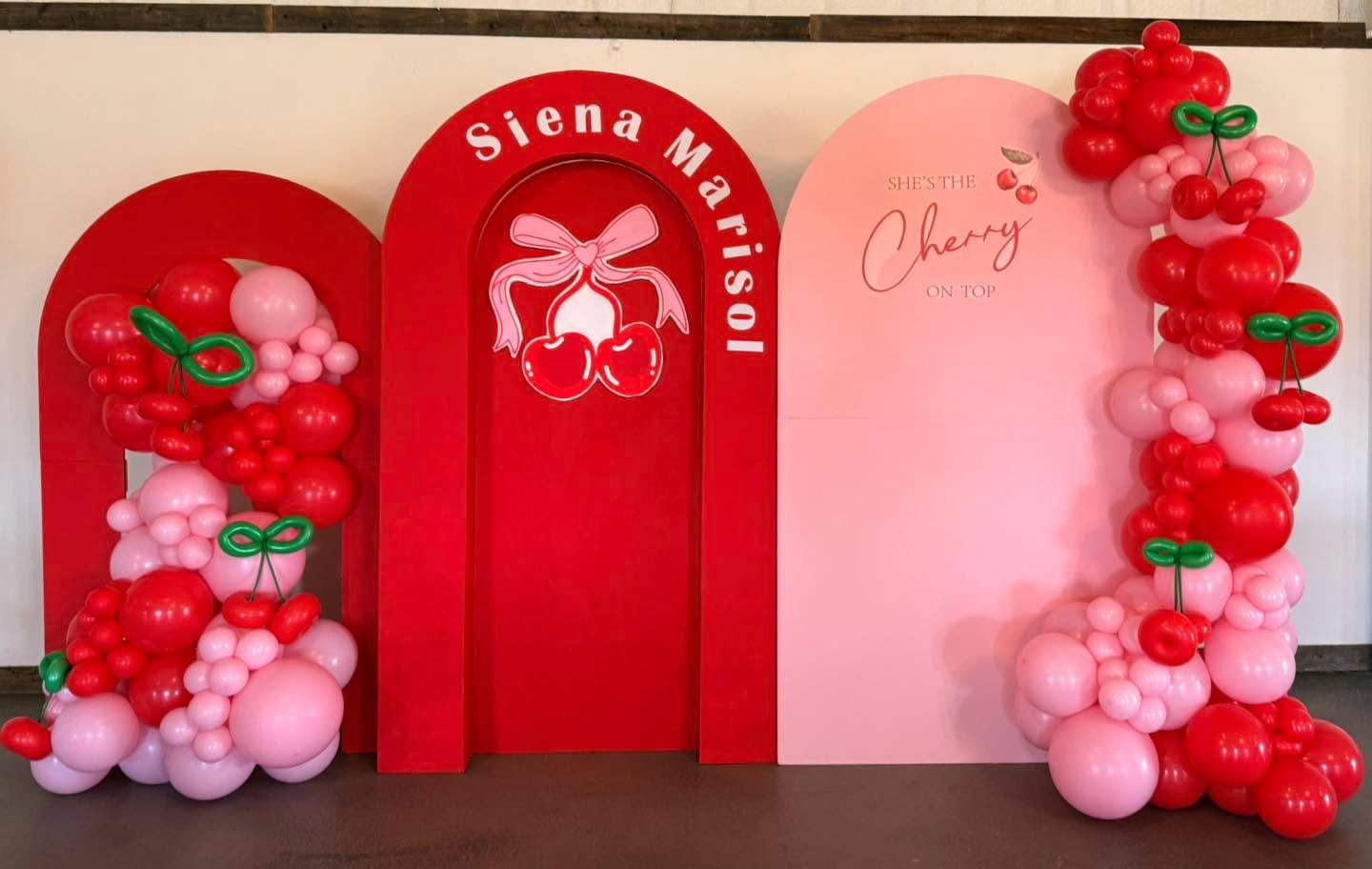 Cherry-themed balloon decoration with red and pink balloons shaped like cherries and leaves, a red backdrop with a cherry graphic and the text "Siena Marisa!", and a pink panel with "She's the Cherry on Top" written on it.