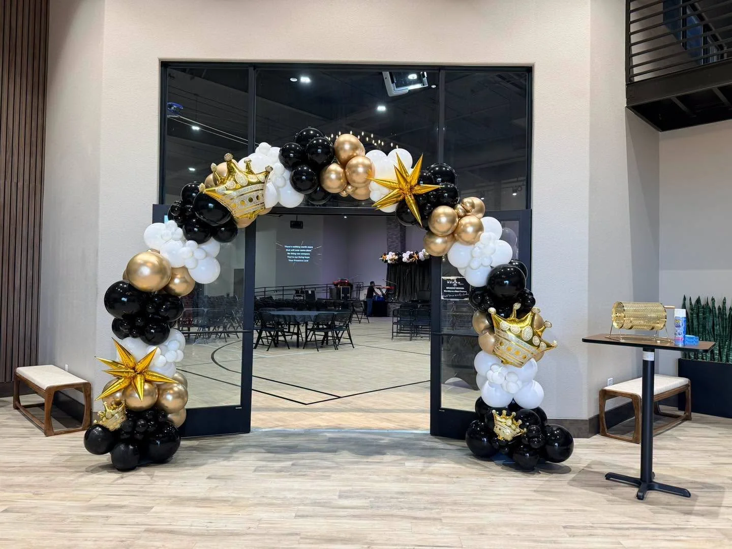 Basketball court decorated with black, white, and gold balloons shaped into a balloon arch with crown and star decorations, set in an indoor gymnasium.